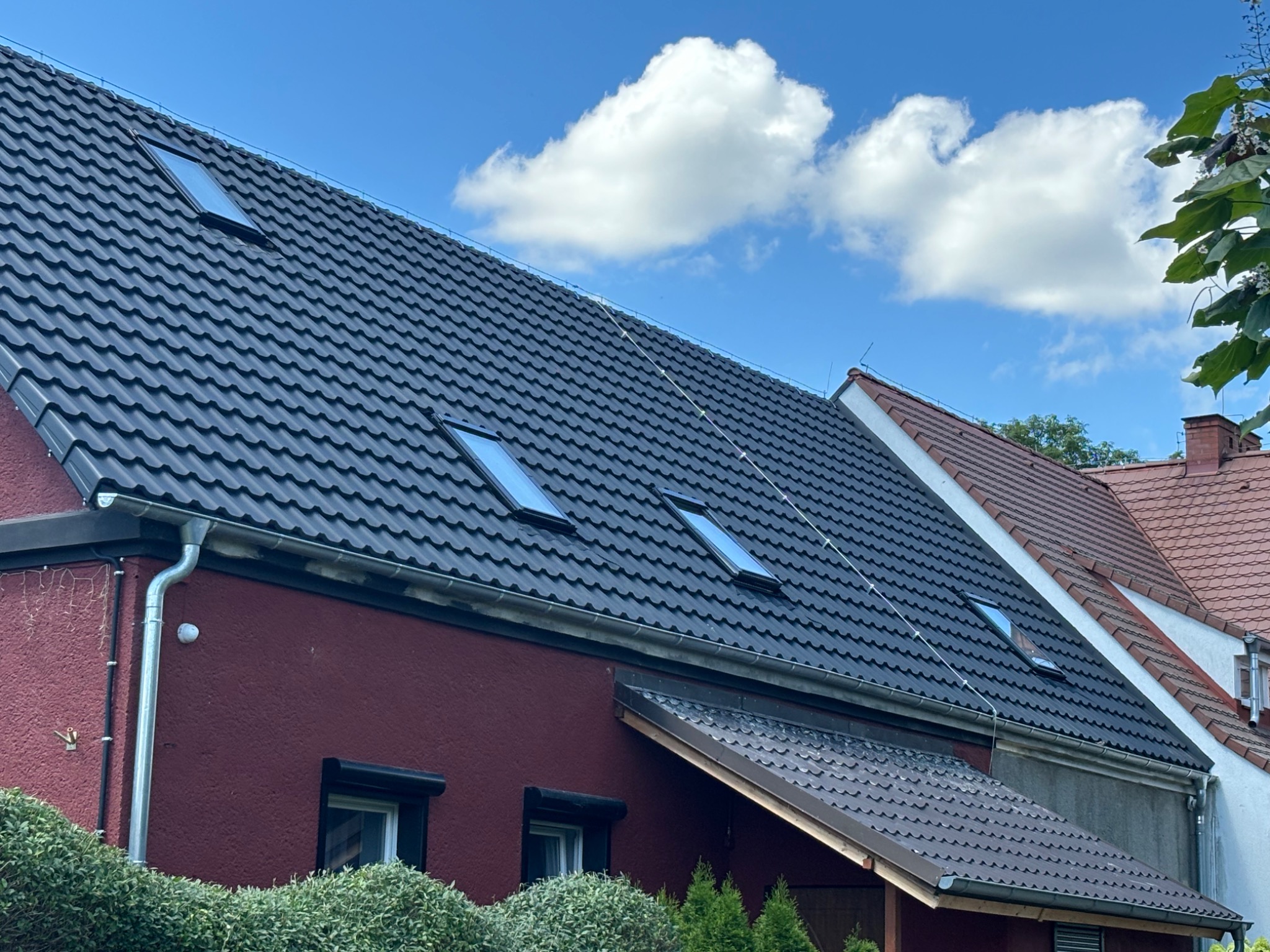 Ciemnoszary dach z dachówki ceramicznej z oknami połaciowymi, widok z boku domu w kolorze bordowym, z widocznymi rynnami i fragmentami innych budynków w tle pod błękitnym niebem z białymi chmurami.