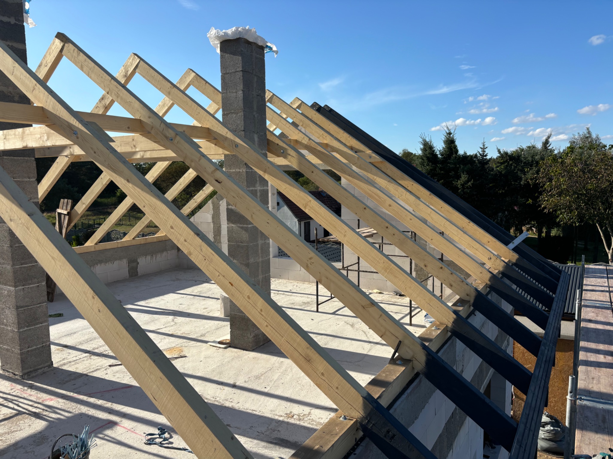 Konstrukcja drewniana więźby dachowej z kominem w trakcie budowy, widok z góry na elementy montażowe i częściowo pokryte czarną membraną dachową, na tle błękitnego nieba.