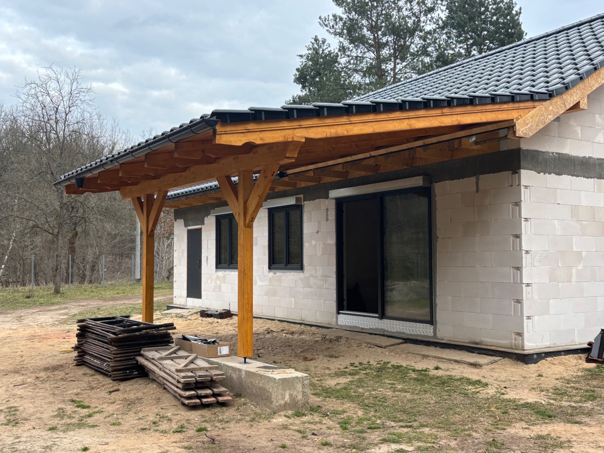 Dom w trakcie budowy z nową drewnianą konstrukcją dachu i częściowo ułożonymi szarymi dachówkami, widoczne przygotowane elementy konstrukcji na ziemi.