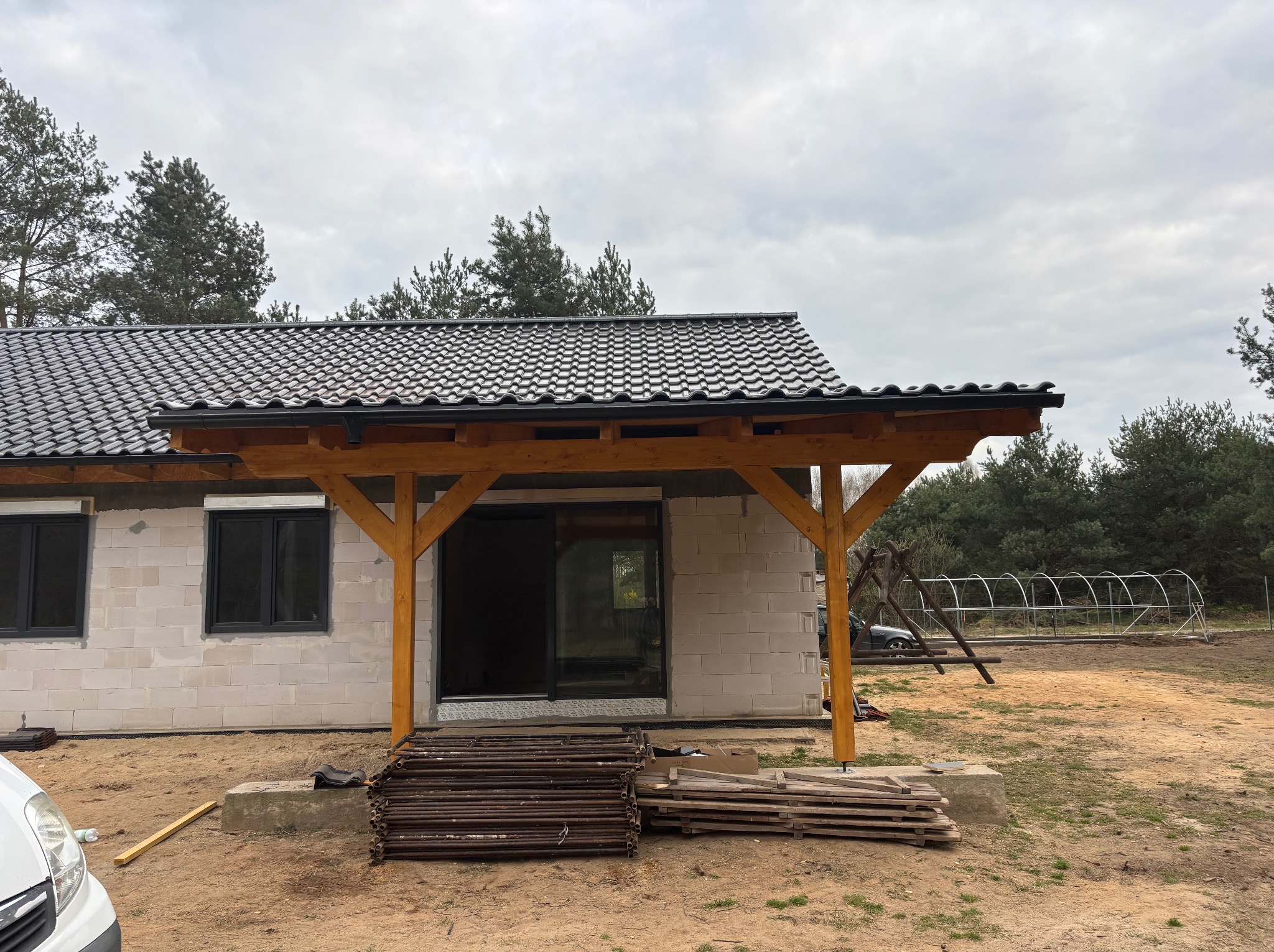 Dom w trakcie budowy z nowym, ciemnoszarym dachem z dachówki ceramicznej i drewnianą konstrukcją zadaszenia nad wejściem, widoczne rusztowania i materiały budowlane na posesji.