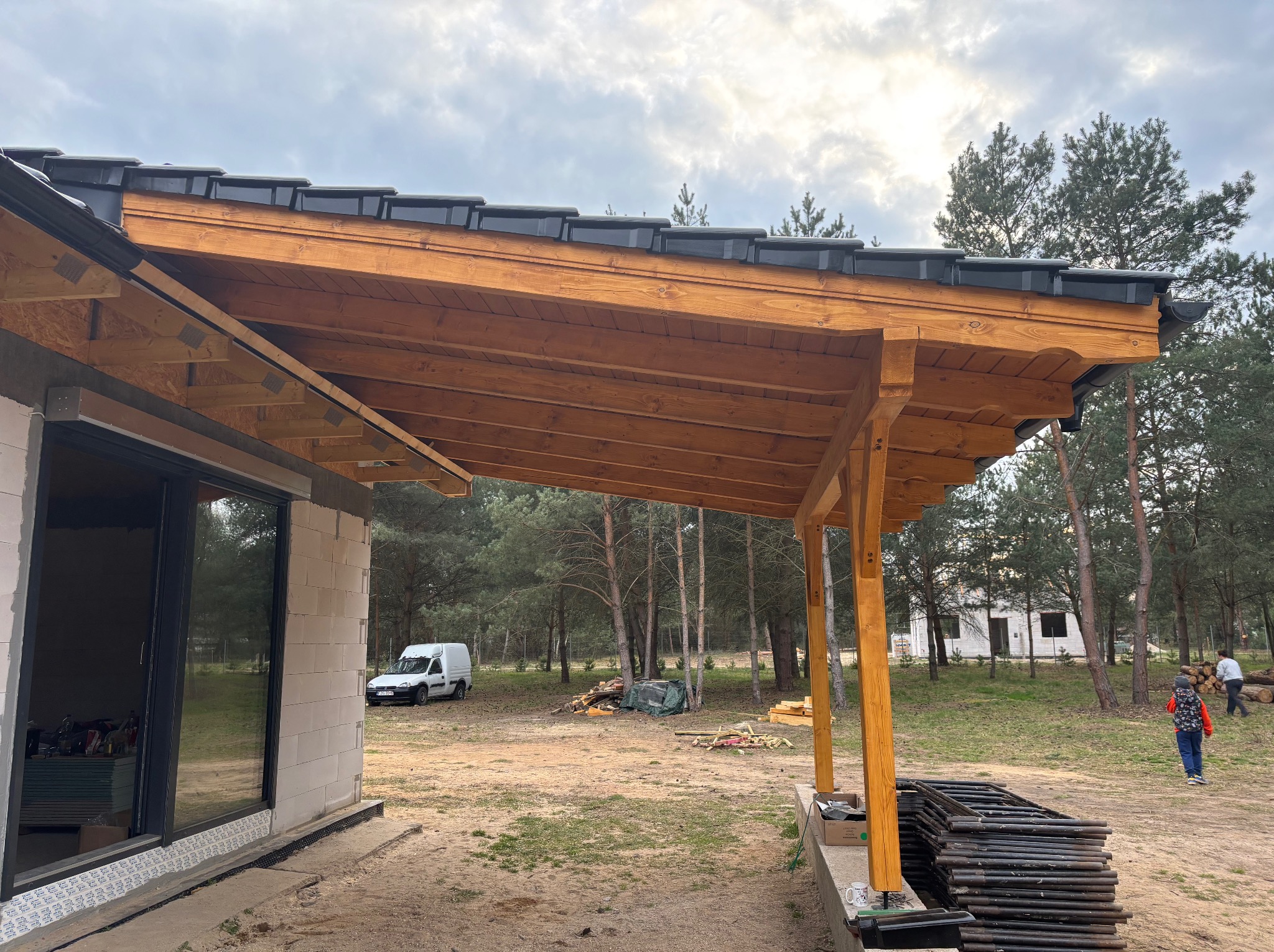 Drewniana konstrukcja dachu nad tarasem w trakcie budowy, widoczne czarne dachówki, w tle las i biały budynek oraz biały samochód dostawczy.
