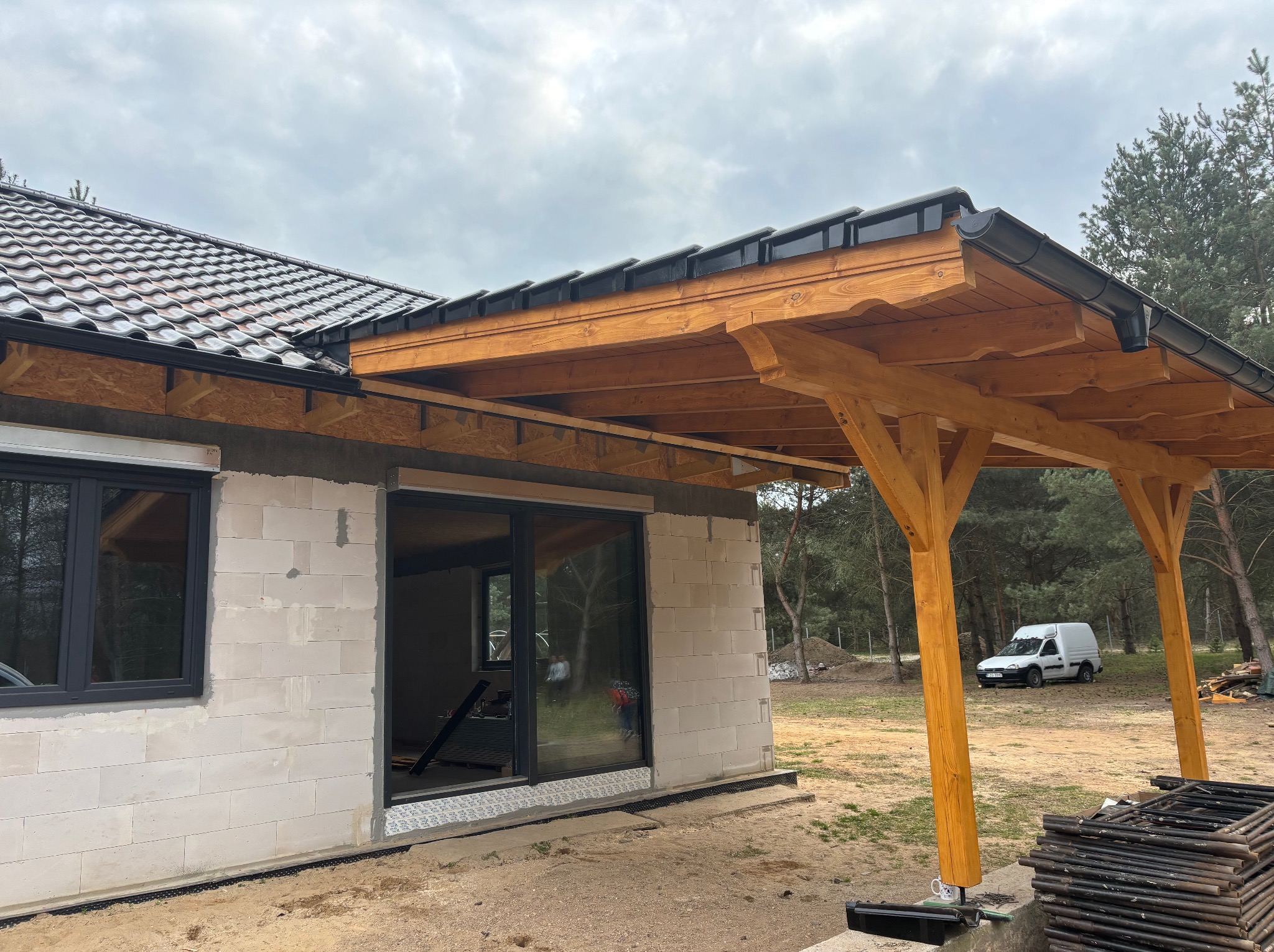 Budynek w trakcie budowy z zamontowaną drewnianą konstrukcją zadaszenia i częściowo położonym dachem z dachówki ceramicznej, widoczne obróbki blacharskie i rynny, na pierwszym planie stos rusztowań.