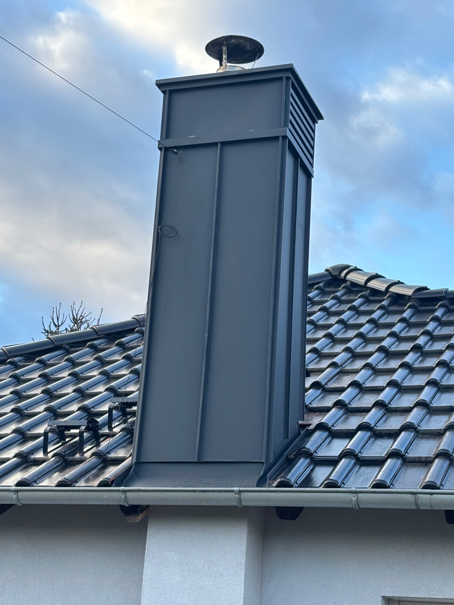 Szary, metalowy komin z daszkiem, wystający ponad ciemny dach z dachówki ceramicznej. Widoczne obróbki blacharskie i fragment rynny. Pochmurne niebo w tle.