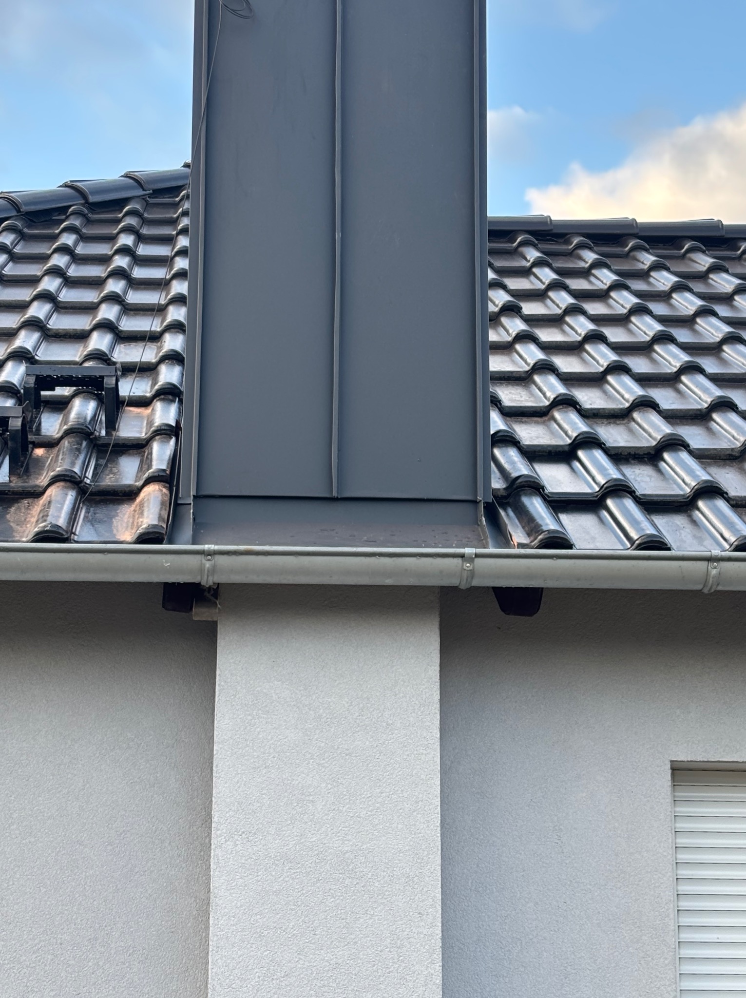 Błyszczący, ciemny dach z ceramicznej dachówki, z szarą obróbką komina z blachy. Widoczna rynna i fragment elewacji budynku z roletą okienną.