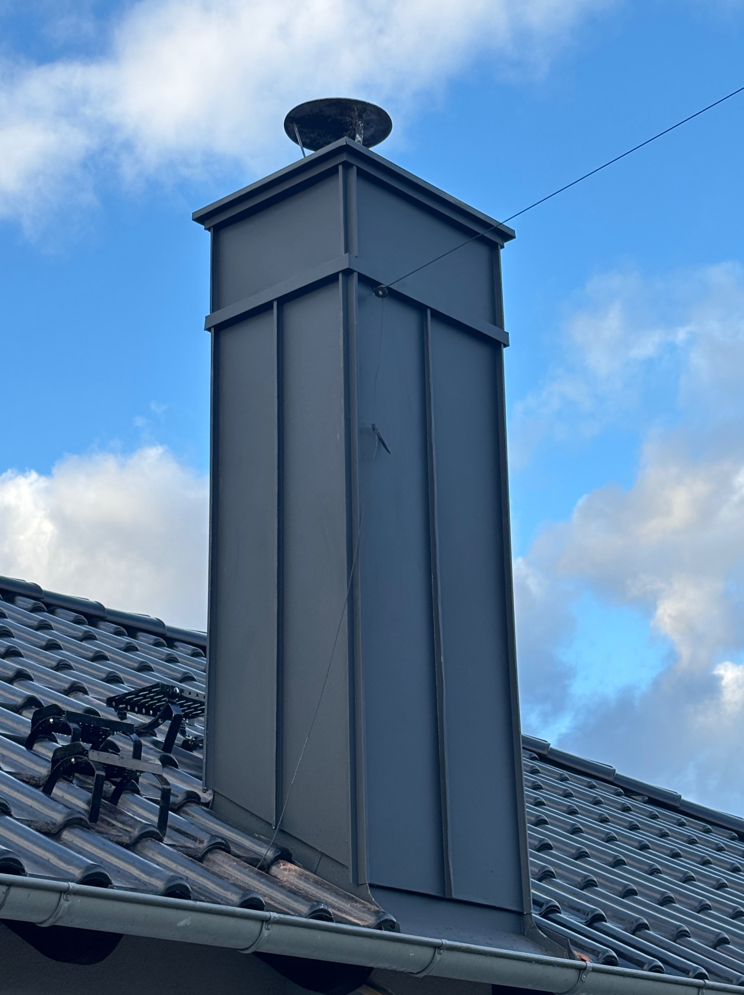 Szary komin z blachy na dachu z dachówki ceramicznej, na tle błękitnego nieba z chmurami. Widoczne zabezpieczenia na dachu i rynna.