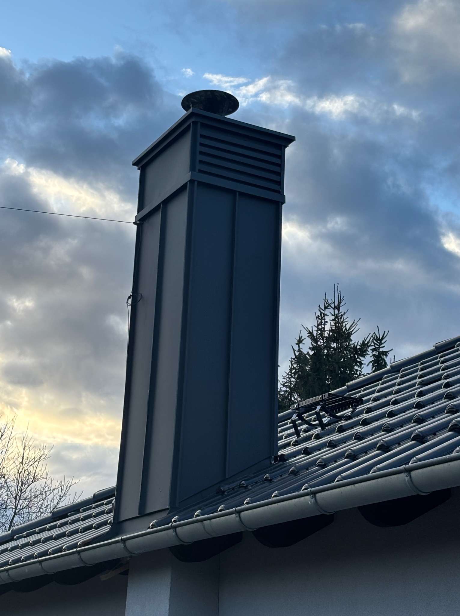 Szary, metalowy komin z daszkiem na tle pochmurnego nieba, widoczny fragment dachu z dachówką i rynną. Na dachu zamontowany stopień kominiarski.