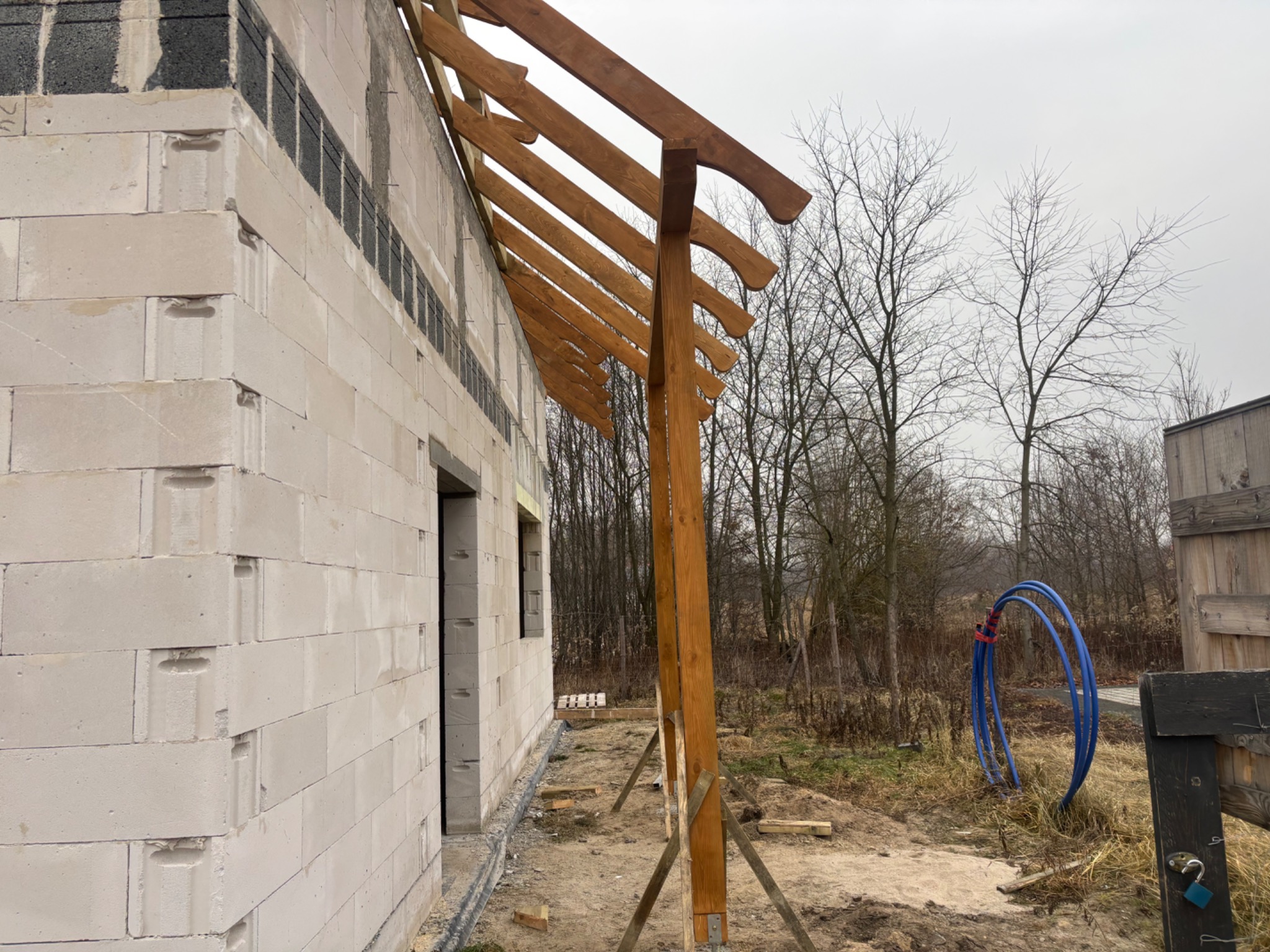 Wznoszenie drewnianej konstrukcji dachu na surowym budynku z betonu komórkowego. Widoczne elementy więźby dachowej i podparcie. W tle drzewa i niebieskie rury.