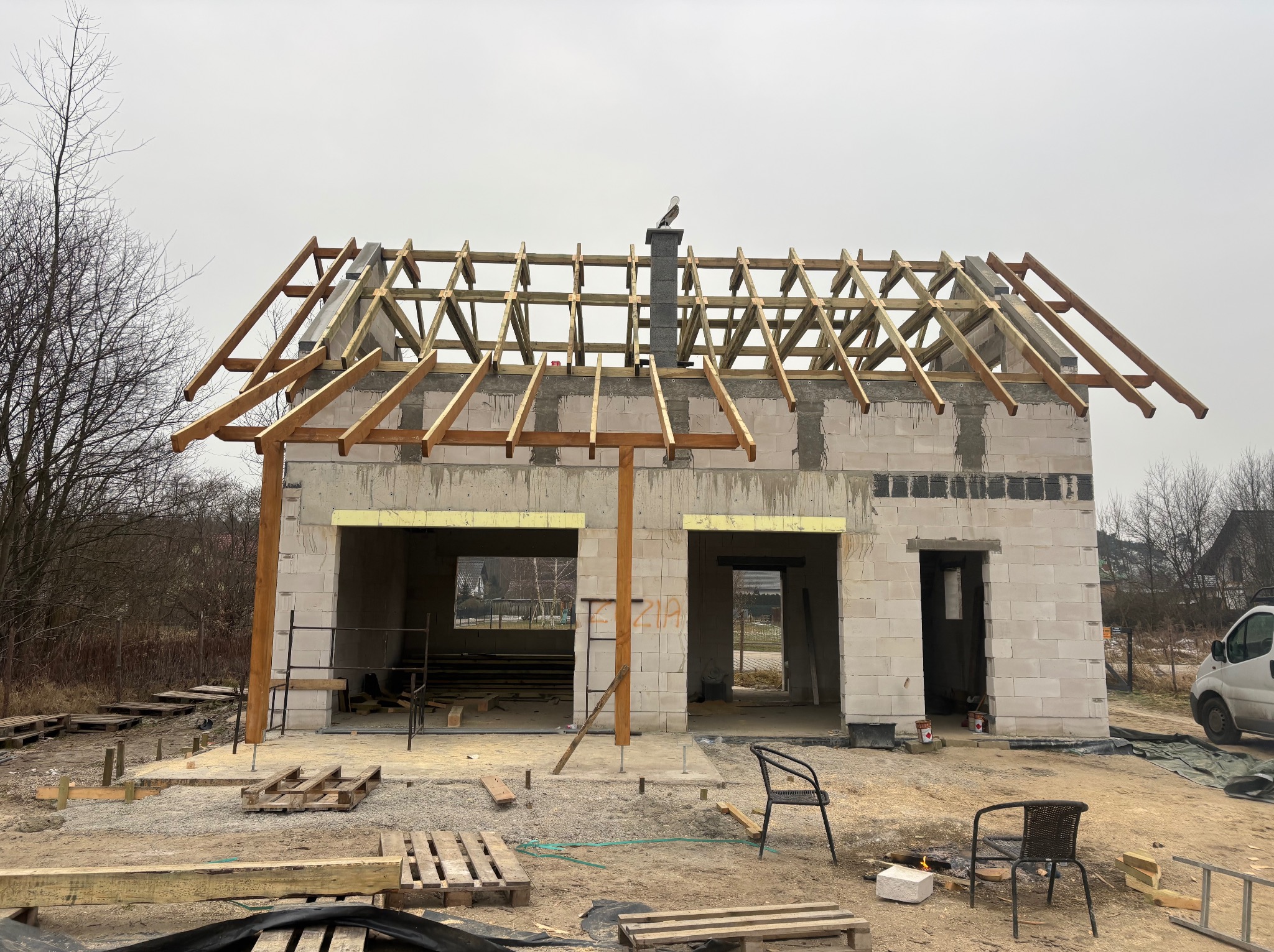 Budowa domu w trakcie: wzniesione ściany z bloczków, drewniana konstrukcja dachu z widocznym kominem. Na kominie siedzi ptak. Widoczne palety i narzędzia budowlane.