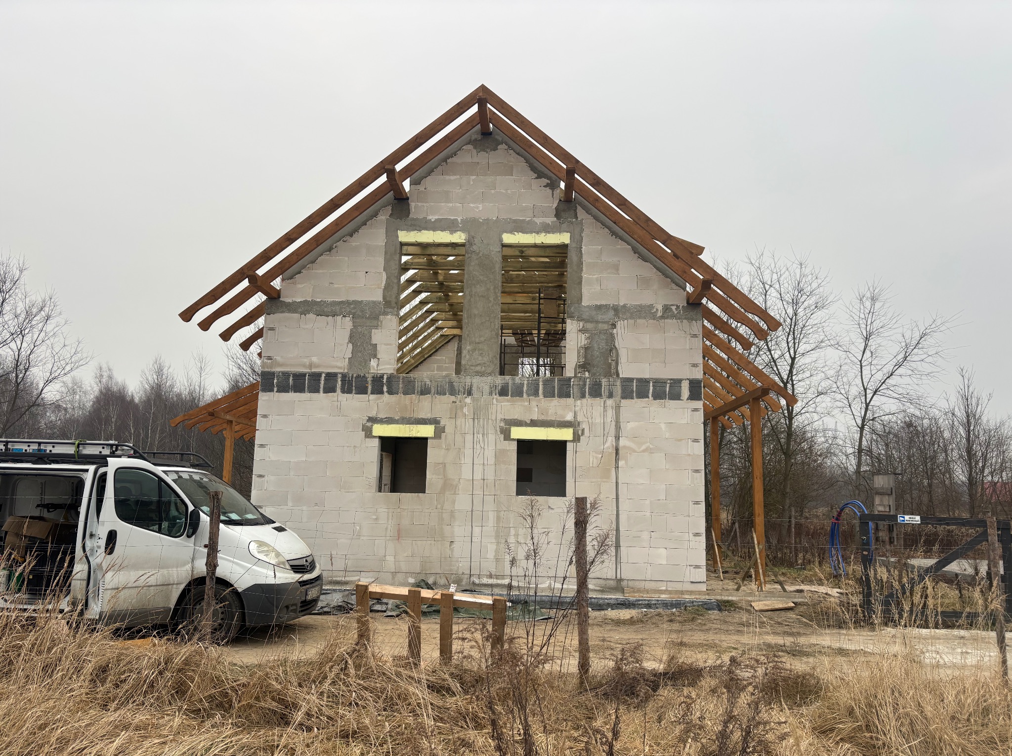 Surowy stan domu z drewnianą konstrukcją dachu w trakcie budowy, z widocznym samochodem dostawczym i elementami rusztowania na pierwszym planie.
