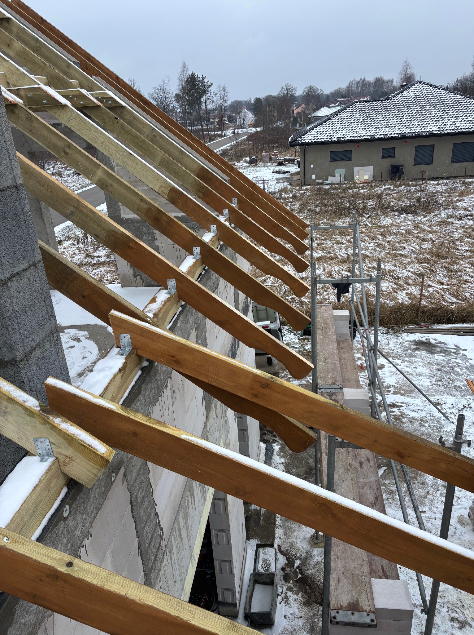 Wznoszenie drewnianej konstrukcji dachu z widocznymi krokwiami i elementami mocującymi, częściowo pokrytymi śniegiem. W tle budynek mieszkalny i zimowy krajobraz.