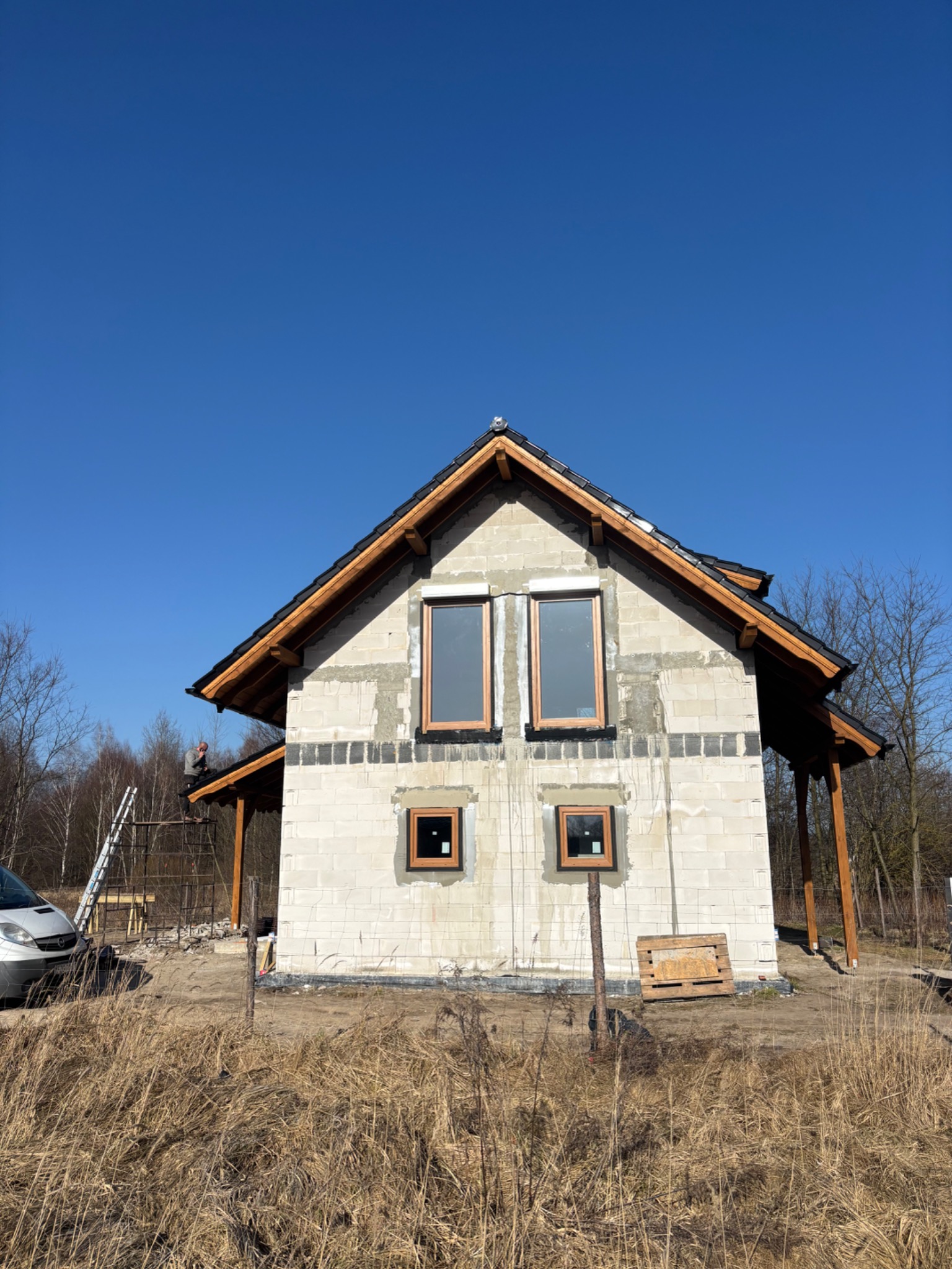 Surowy dom z pustaków z drewnianą konstrukcją dachu i oknami, w trakcie budowy. Na rusztowaniu widoczny pracownik. W tle błękitne niebo i drzewa.