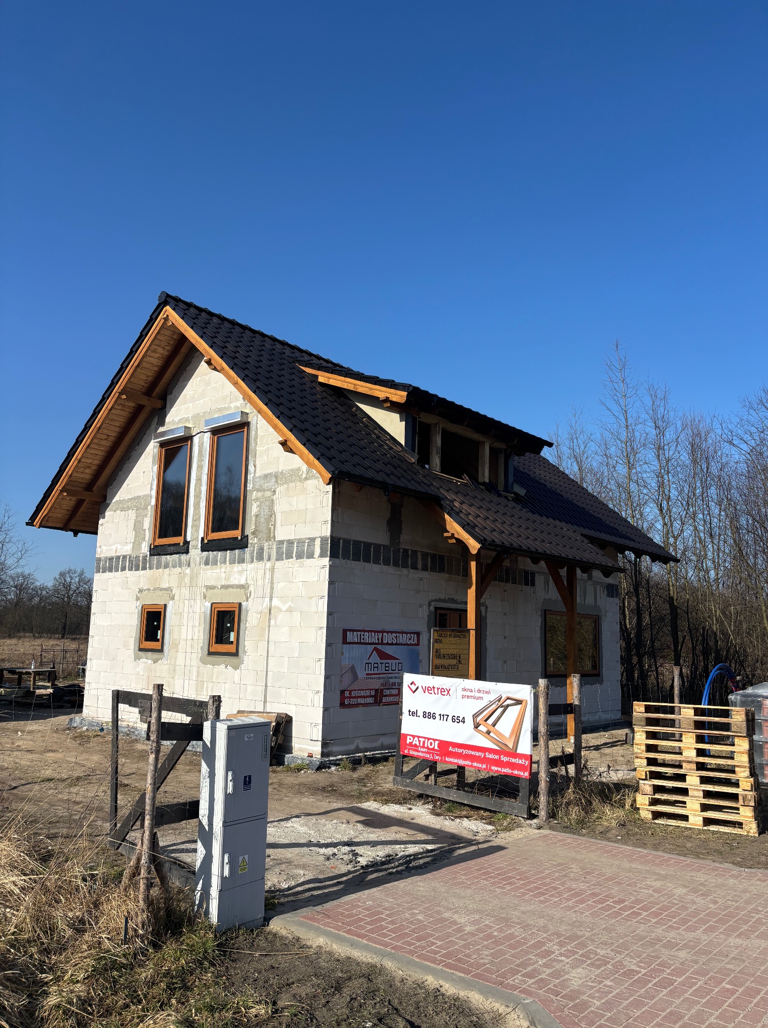 Dom w trakcie budowy z czarnym dachem i drewnianą konstrukcją. Widoczne okna z brązowymi ramami i reklamy materiałów budowlanych. Błękitne niebo w tle.