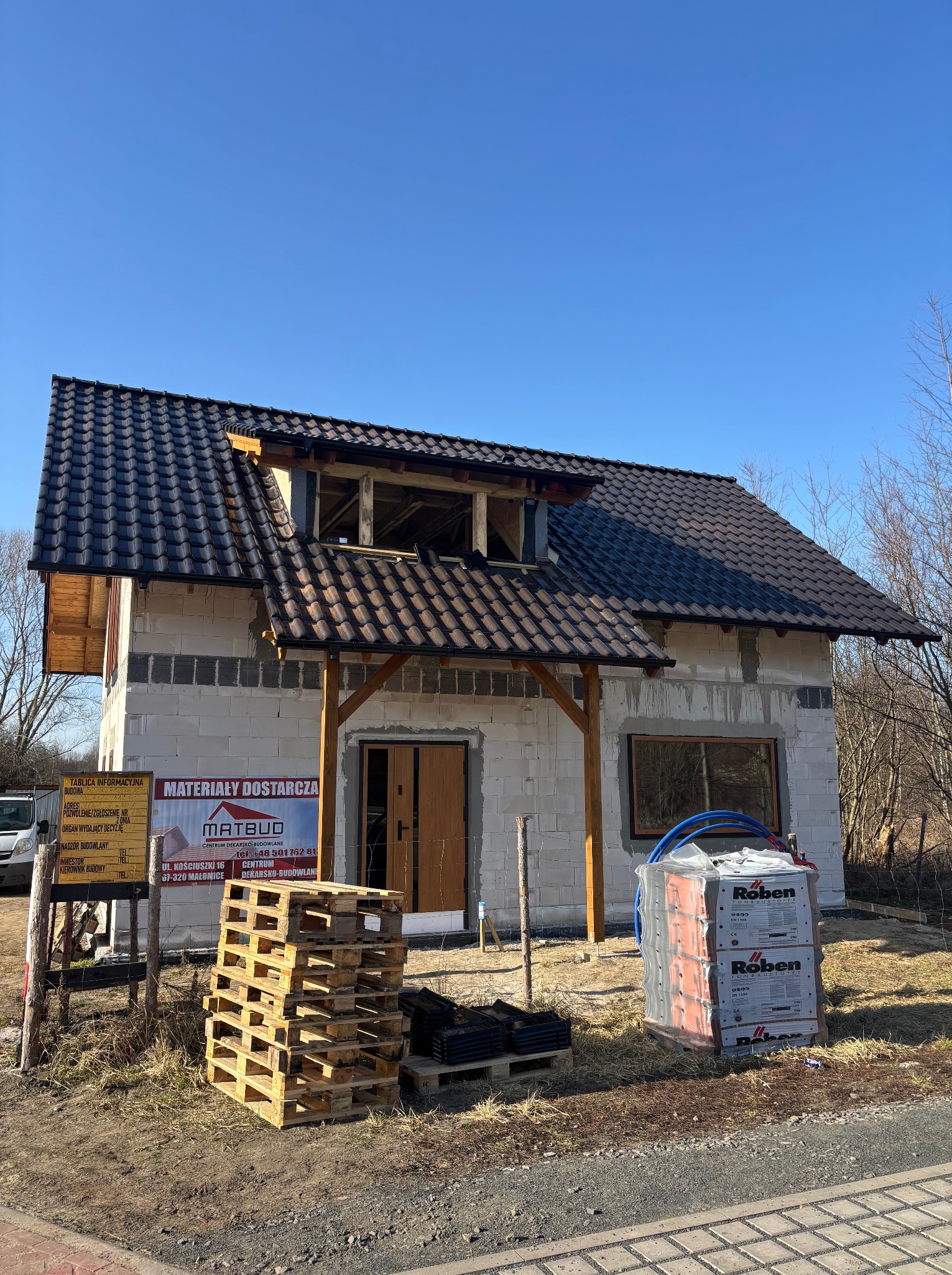 Dom w trakcie budowy z częściowo położonym, ciemnym dachem z dachówki ceramicznej. Widoczne palety i materiały budowlane. Tablica informacyjna budowy.