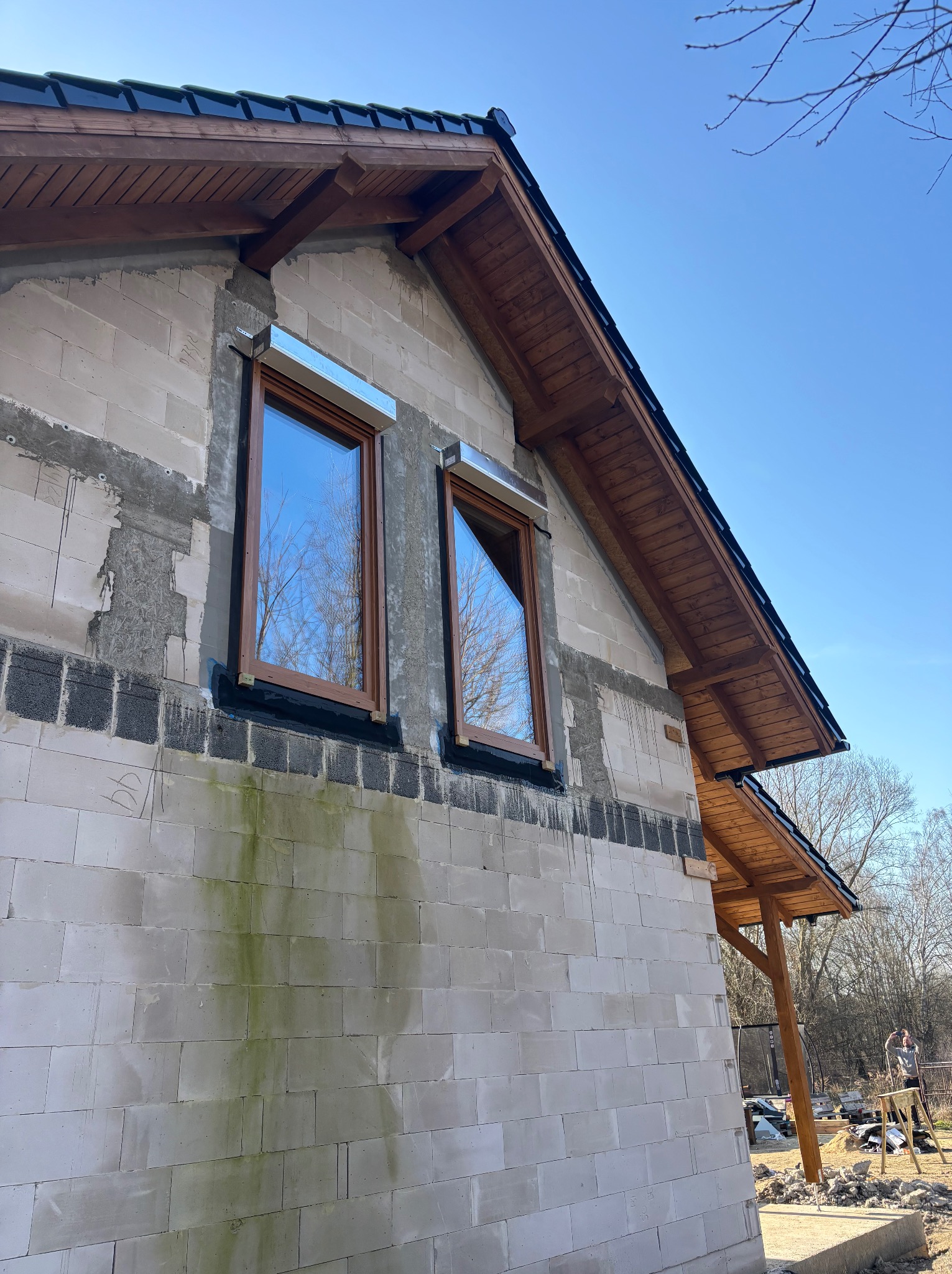 Surowy stan budynku z oknami i fragmentem dachu z drewnianą podbitką, widok z dołu na tle błękitnego nieba. Widoczne ślady wilgoci na ścianie.