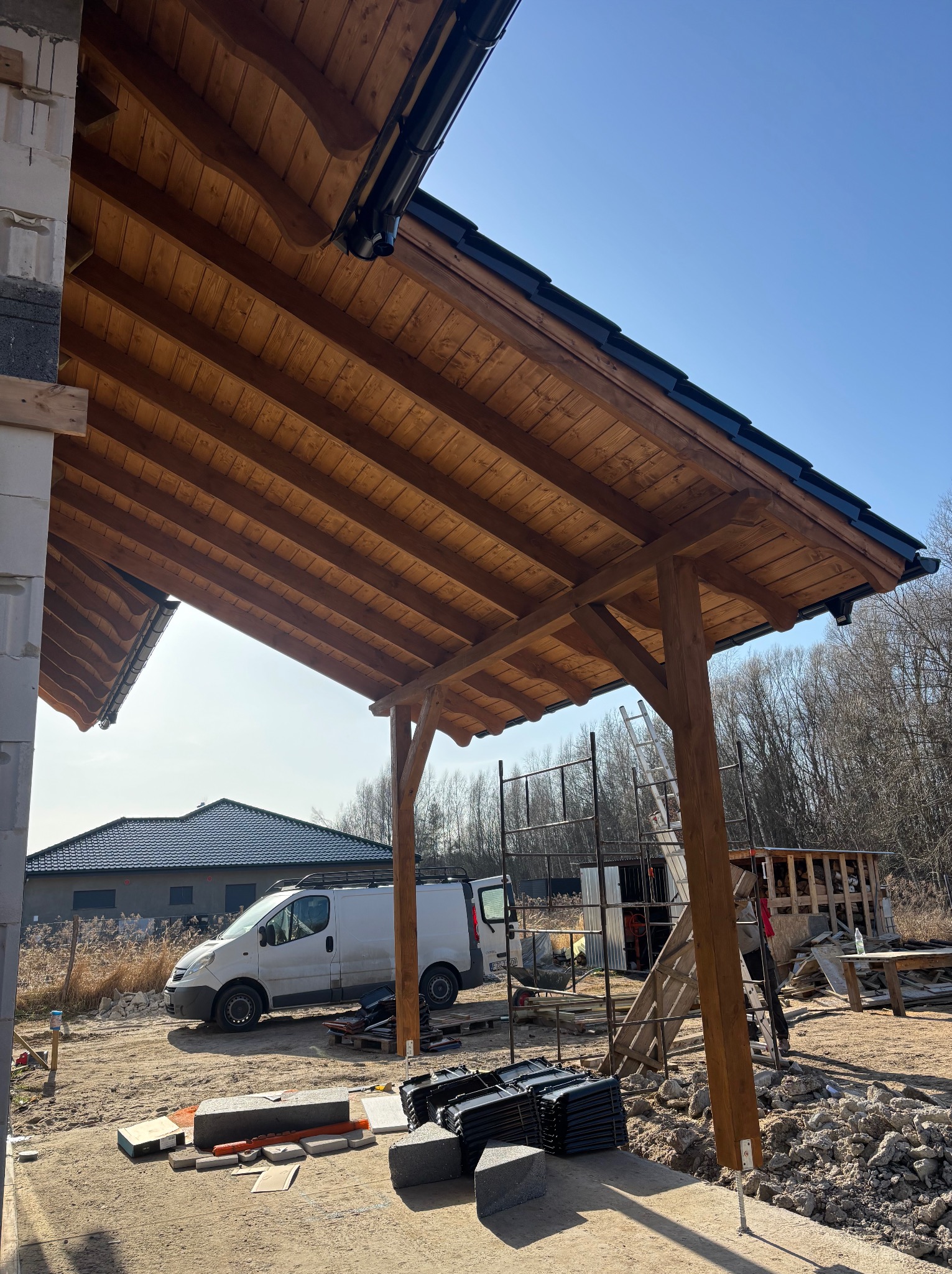 Drewniana konstrukcja z elementami dachu w trakcie budowy, widoczne rusztowania i materiały budowlane. W tle dom i samochód dostawczy.