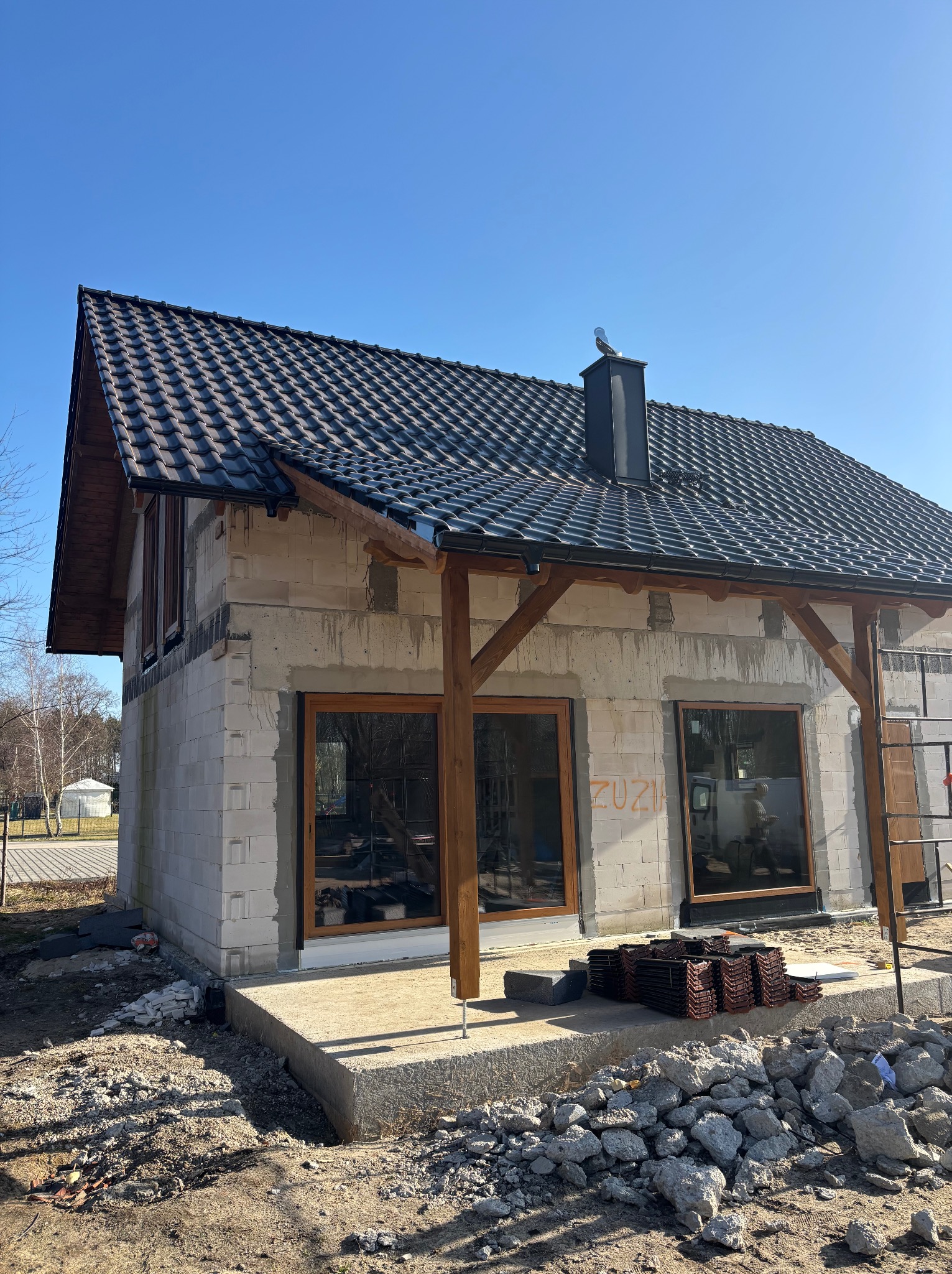 Dom w trakcie budowy z ciemną dachówką i drewnianą konstrukcją werandy. Na dachu widoczny komin z ozdobną figurką. Wokół budynku gruz i materiały budowlane.