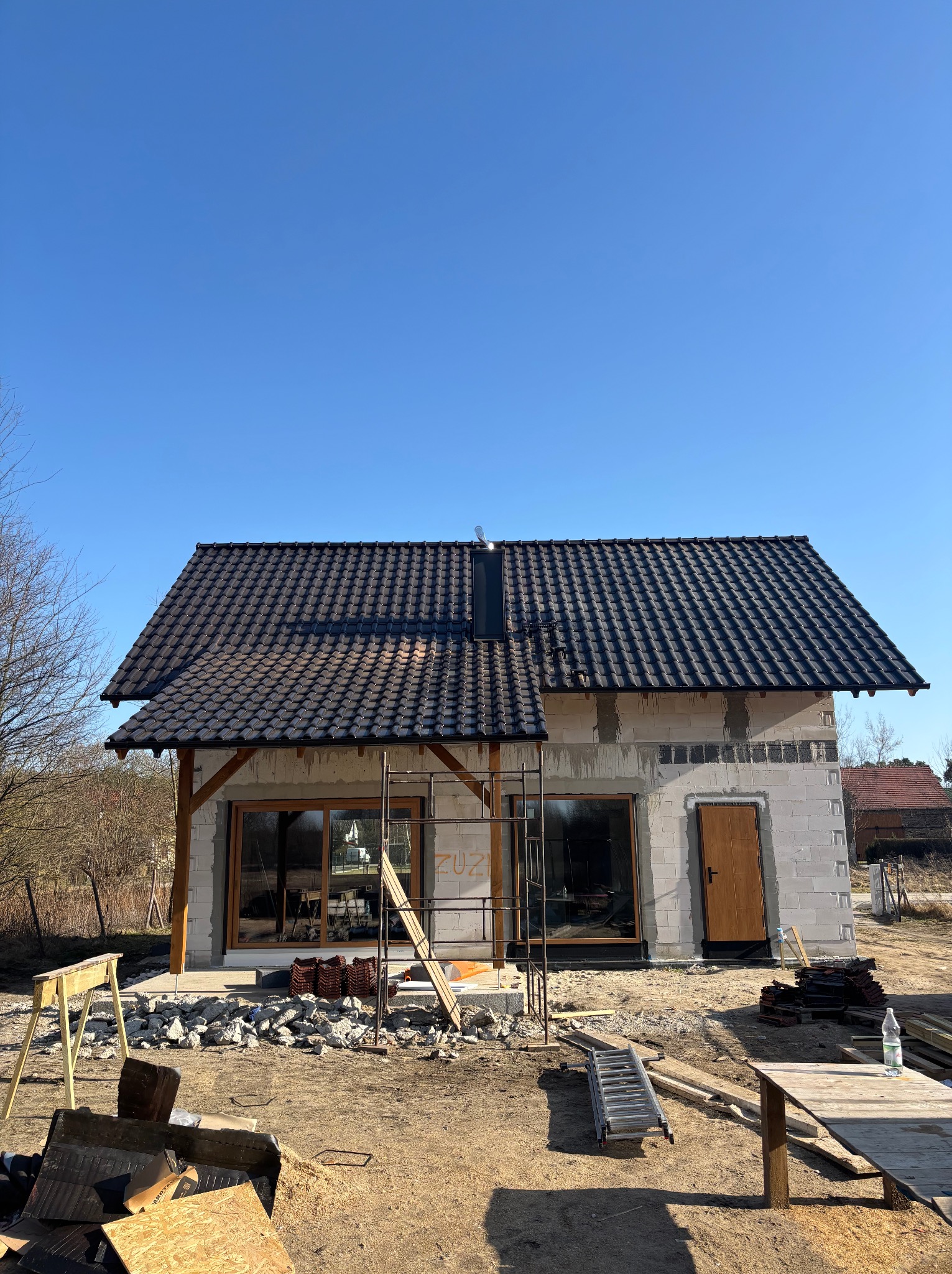 Dom jednorodzinny w trakcie budowy, z ciemną dachówką ceramiczną i widoczną konstrukcją więźby. Na pierwszym planie materiały budowlane i rusztowania.