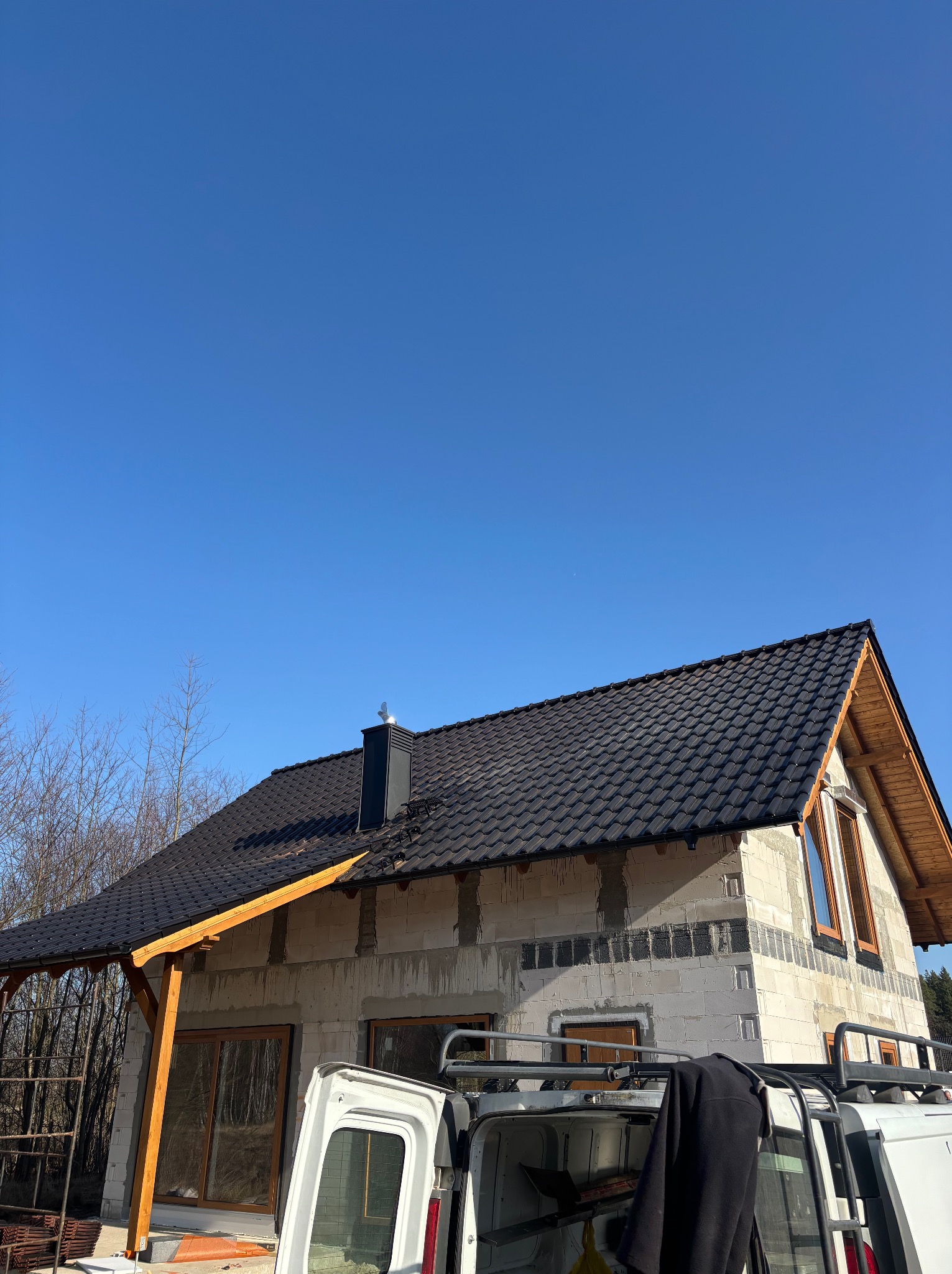 Dom w trakcie budowy z czarnym dachem i drewnianymi elementami wykończeniowymi, widoczny zza białego samochodu dostawczego w słoneczny dzień.