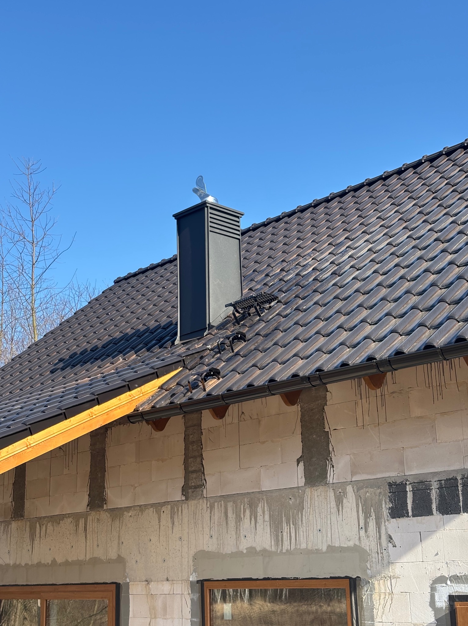 Nowy, brązowy dach z dachówki ceramicznej, czarny komin z ozdobną wiatrowskazą, zamontowane rynny, widoczna ściana budynku w trakcie budowy.