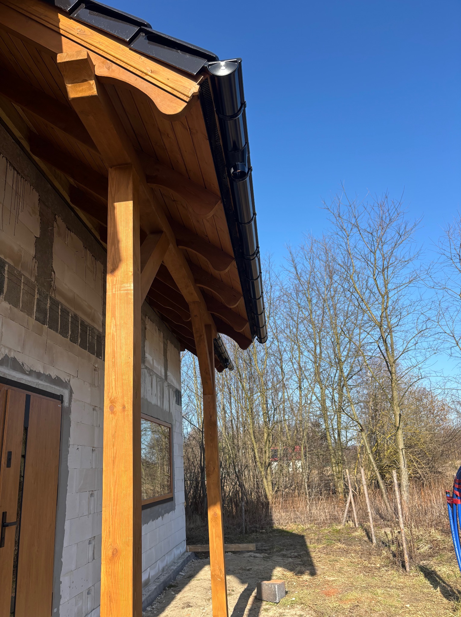 Nowoczesny, czarny system rynnowy zamontowany na drewnianej konstrukcji zadaszenia, widok z dołu na tle błękitnego nieba i drzew bez liści.