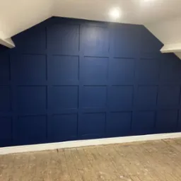 Ściana w pokoju na poddaszu, pomalowana na granatowo z dekoracyjnymi panelami w kształcie kwadratów, biała listwa przypodłogowa, surowa drewniana podłoga.