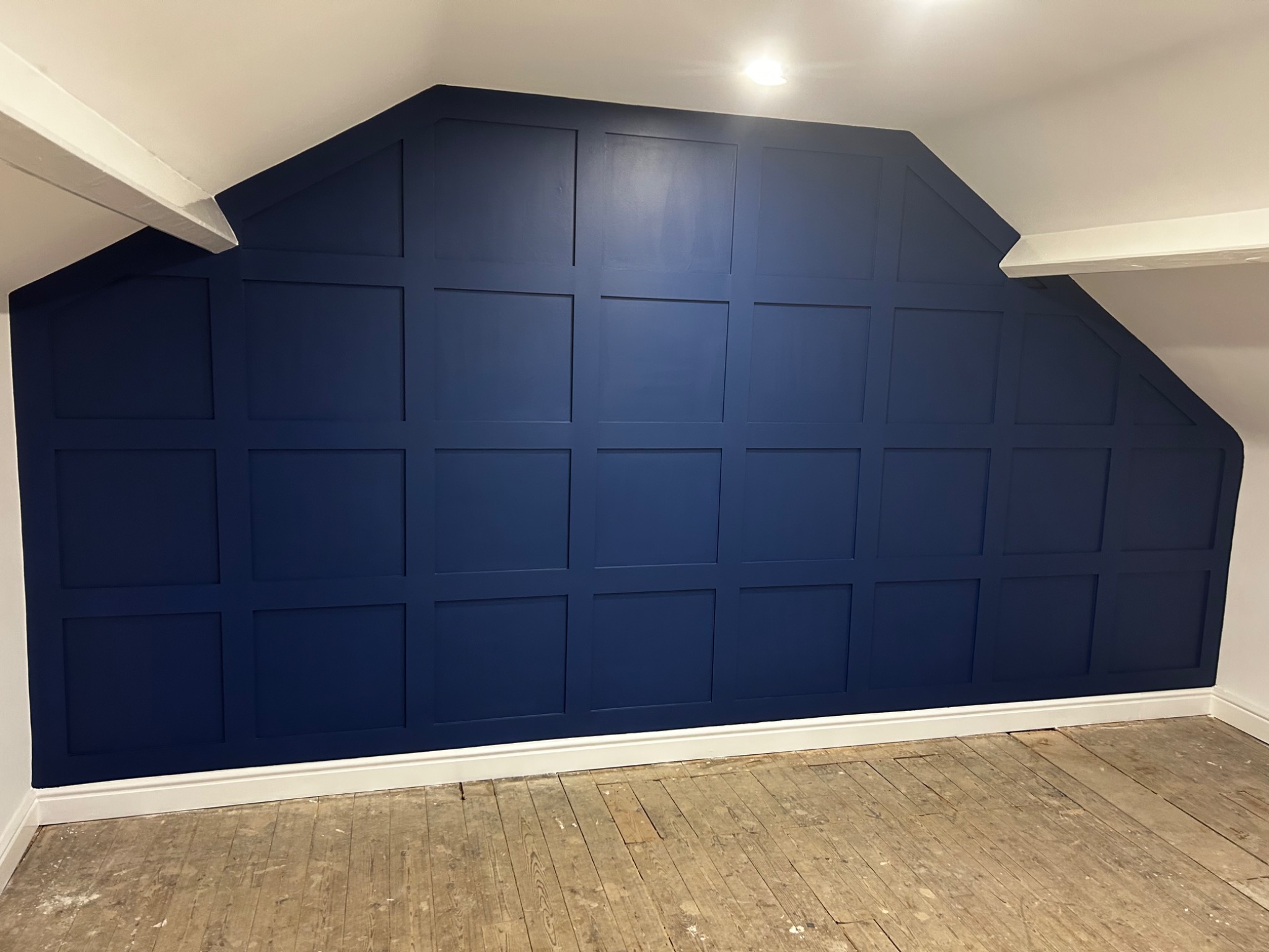 Ściana w pokoju na poddaszu, pomalowana na granatowo z dekoracyjnymi panelami w kształcie kwadratów, biała listwa przypodłogowa, surowa drewniana podłoga.