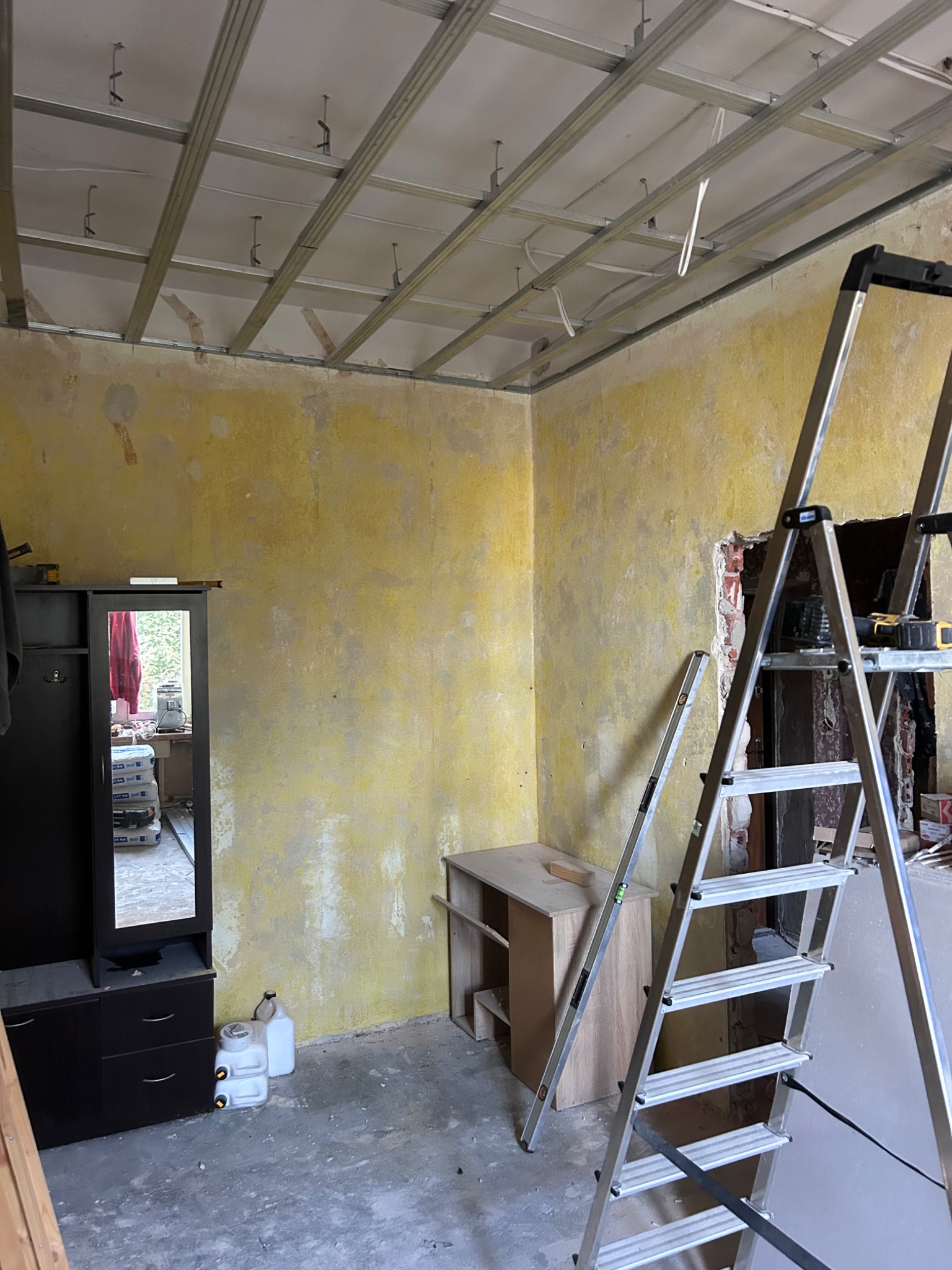Wnętrze w trakcie remontu z widoczną metalową konstrukcją sufitu podwieszanego, żółtą ścianą z pozostałościami farby, drabiną rozstawną i meblami w tle.