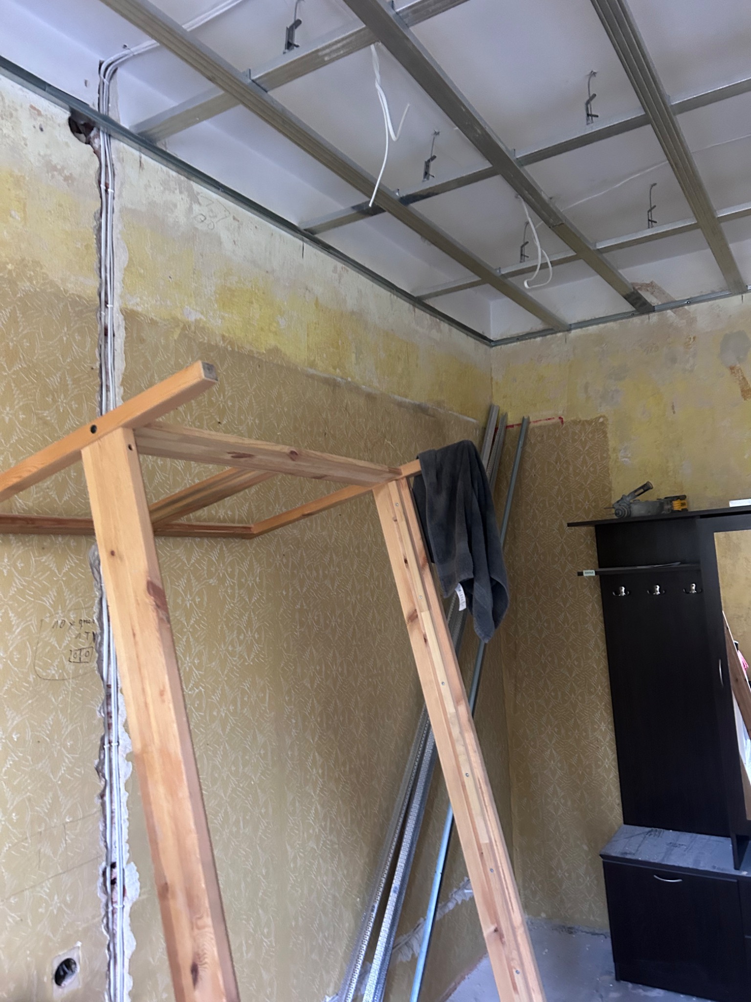 Metalowa konstrukcja sufitu podwieszanego z płyt kartonowo-gipsowych zamontowana w pomieszczeniu z widocznymi starymi tapetami na ścianach i wystającymi przewodami elektrycznymi, z drewnianą...