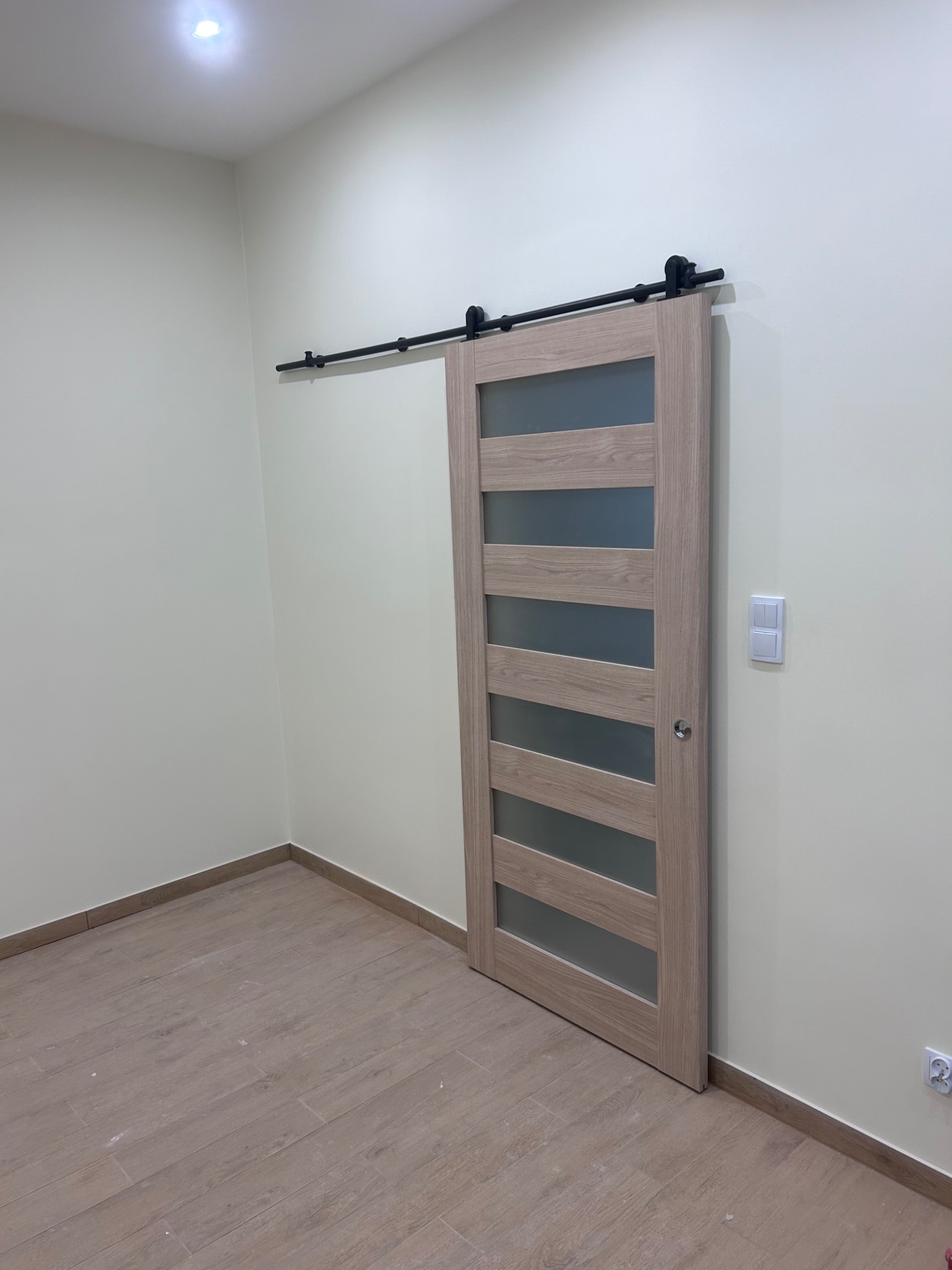 Zamknięte przesuwne drzwi w stylu stodoły z pięcioma matowymi szybami, zamontowane na czarnej szynie w jasnym pokoju z panelami podłogowymi.