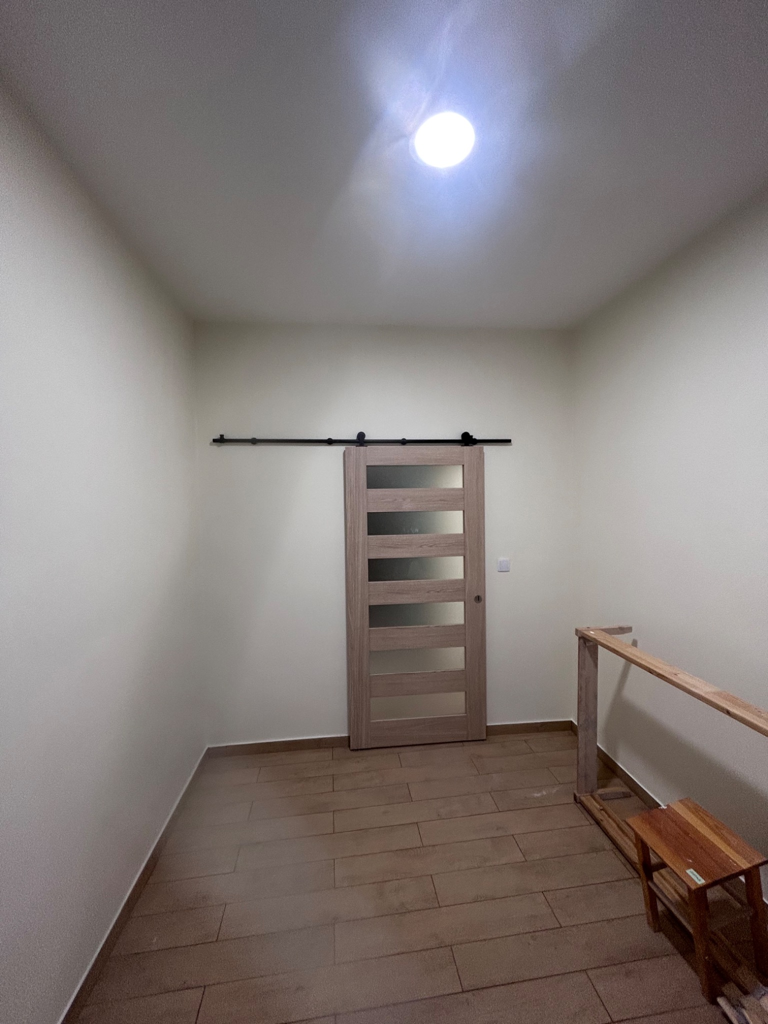 Zamontowane przesuwne drzwi z szybami w jasnej ościeżnicy, zawieszone na czarnym szynowym systemie, z widoczną podłogą z paneli imitujących drewno i fragmentem drewnianej konstrukcji w surowym stanie.