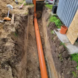 Pomarańczowa rura kanalizacyjna ułożona w wykopie ziemnym, widoczne narzędzia i materiały budowlane w tle.