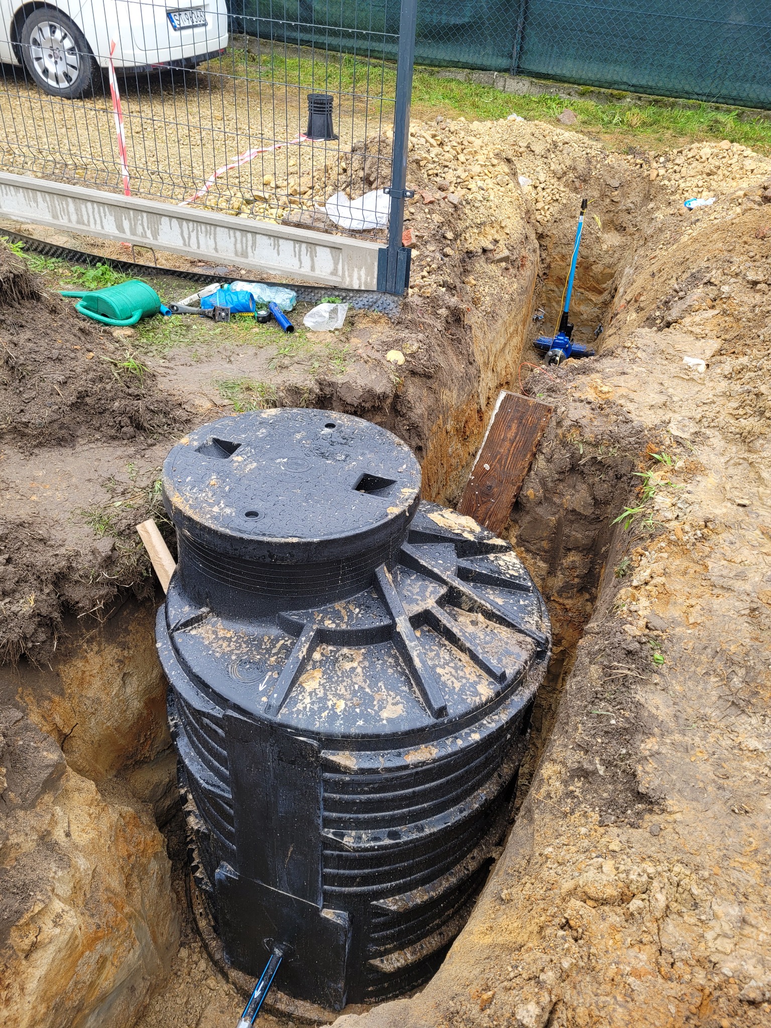 Czarny zbiornik bezodpływowy, prawdopodobnie szambo, umieszczony w wykopie w Jawiszowicach, z widocznymi narzędziami i elementami instalacji w tle.