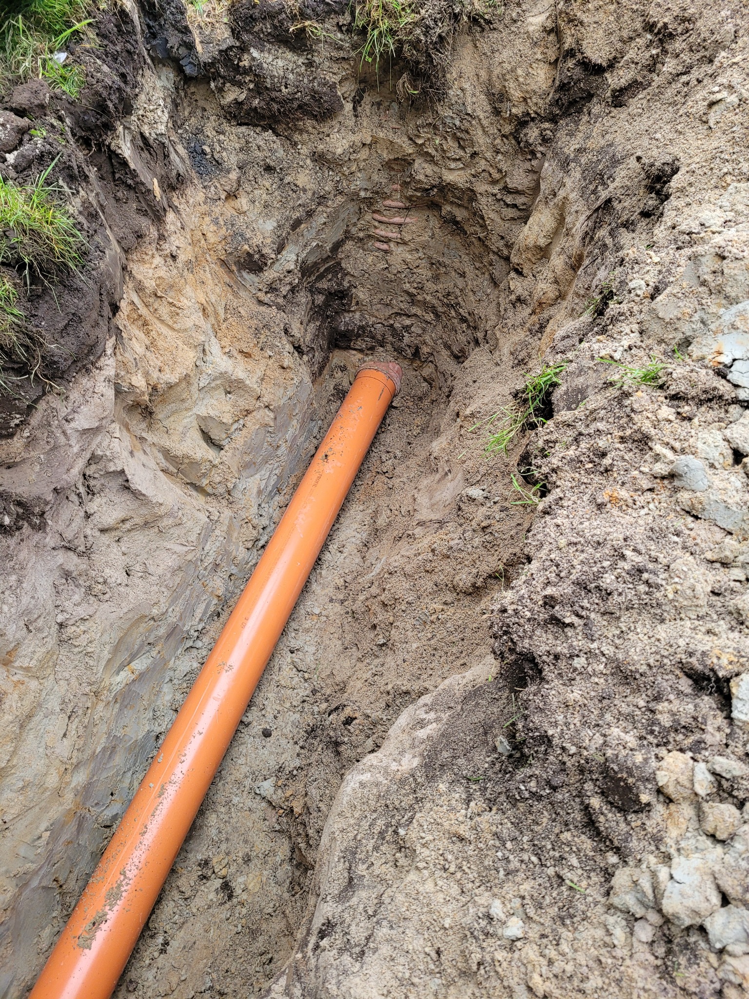 Pomarańczowa rura ułożona w wykopie ziemnym z widoczną strukturą warstw gleby, trawa na krawędzi wykopu.