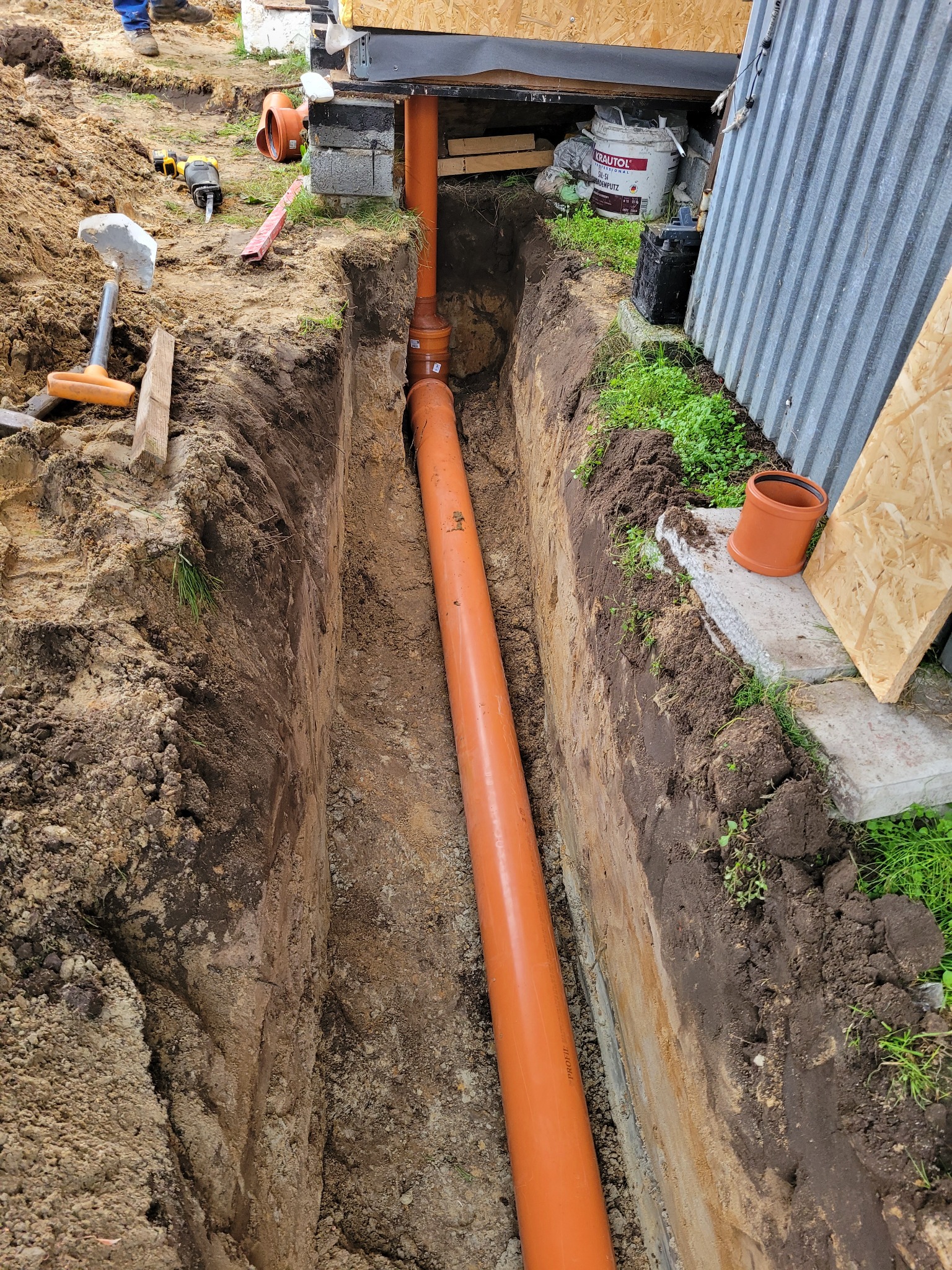 Pomarańczowa rura kanalizacyjna ułożona w wykopie ziemnym, widoczne narzędzia i materiały budowlane w tle.