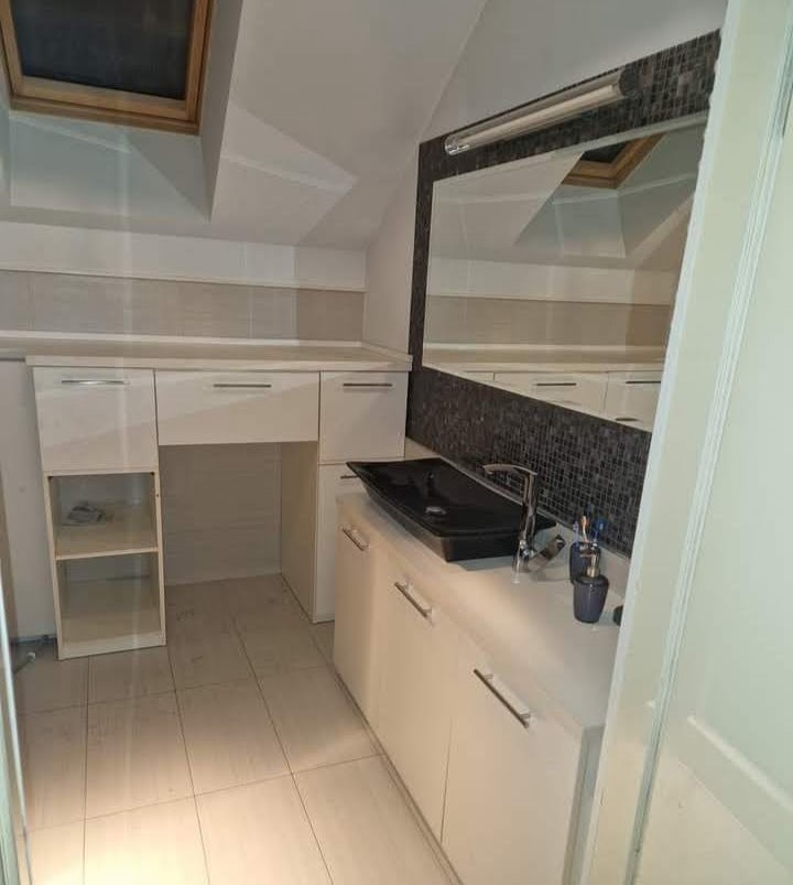 Wykończona łazienka na poddaszu: czarna, prostokątna umywalka nablatowa na białej szafce z szufladami, duże lustro z mozaikową ramą, płytki w odcieniach beżu i szarości, okno dachowe.