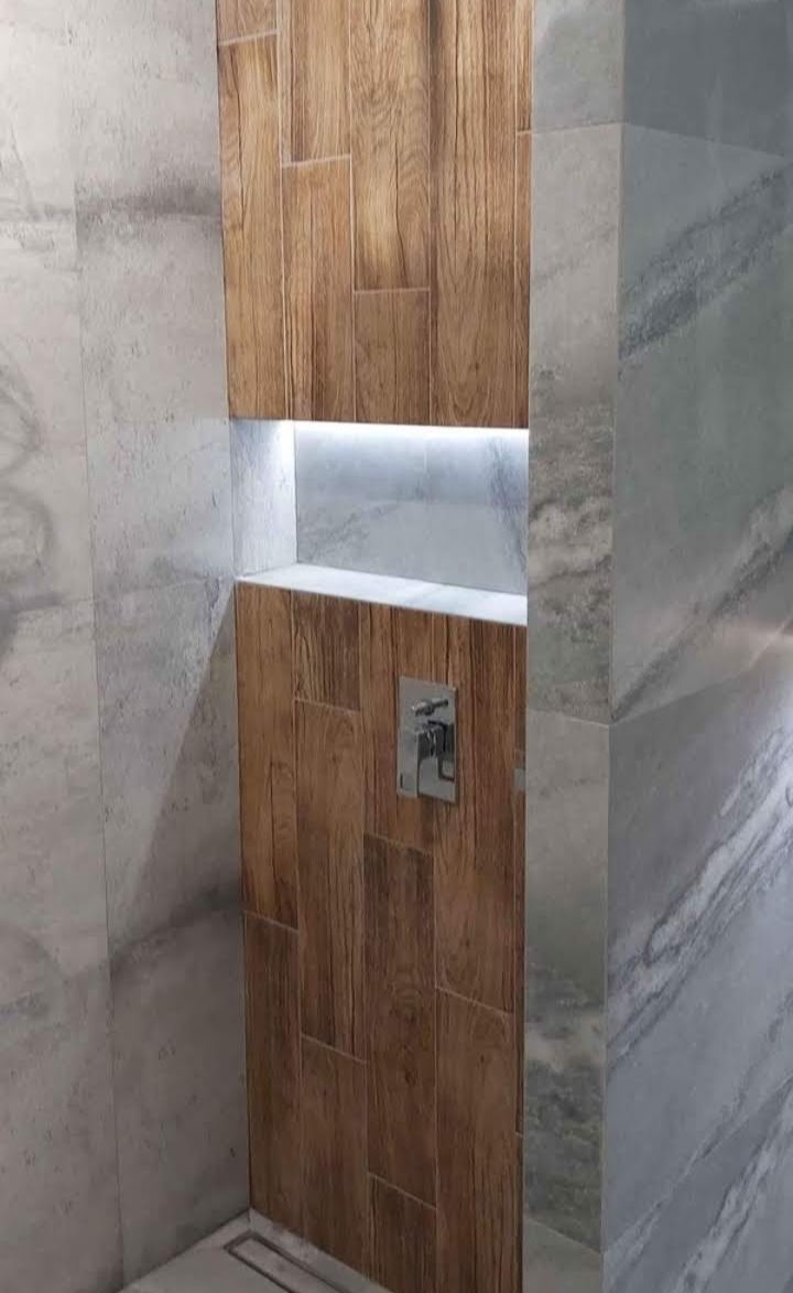 Wykończona wnęka prysznicowa z drewnopodobnymi płytkami, podświetlona listwą LED, z baterią natryskową i odpływem liniowym w podłodze, otoczona szarymi płytkami imitującymi marmur.