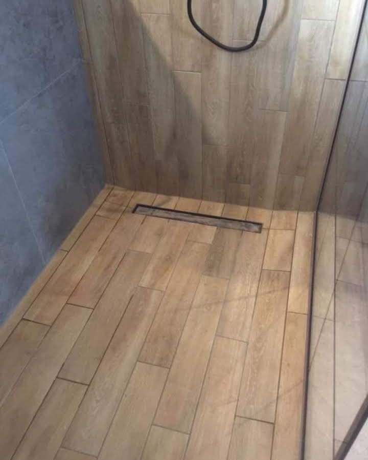 Kabina prysznicowa z drewnopodobnymi płytkami ułożonymi w jodełkę i liniowym odpływem, widoczna szklana ścianka.