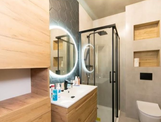 Nowoczesna łazienka z drewnianymi szafkami, okrągłym lustrem z podświetleniem LED, szarą mozaiką heksagonalną i kabiną prysznicową z czarnymi ramami.