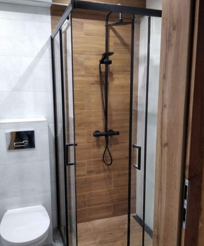 Nowoczesna kabina prysznicowa z czarnymi profilami i deszczownicą, obok toaleta z podtynkowym systemem spłukiwania, ściany wyłożone płytkami imitującymi drewno i białymi płytkami.