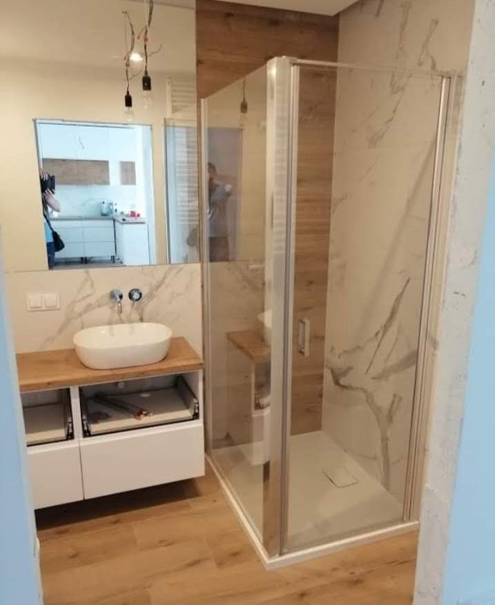 Nowoczesna łazienka z kabiną prysznicową ze szklanymi drzwiami, umywalką nablatową na drewnianym blacie i drewnianą podłogą, widok z boku.
