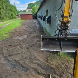 Żółta koparka z łyżką przy budynku z szarym tynkiem i rzędem czarnych okien, przygotowująca teren podłoża z widocznymi śladami opon i błotem.