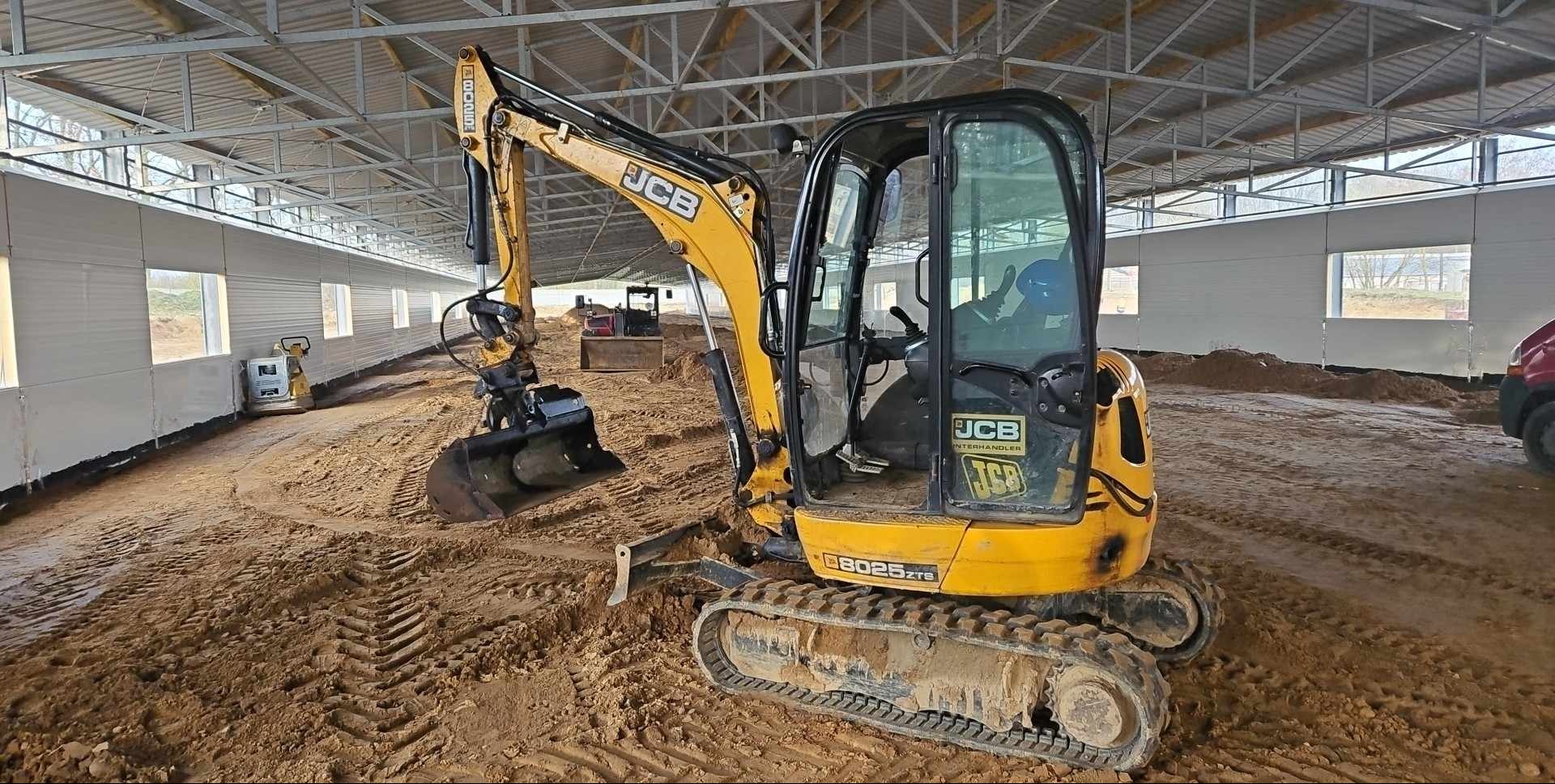 Żółta mini koparka JCB 8025 ZTS na placu budowy wewnątrz hali, widoczne ślady opon w piasku, w tle ubijarka i inna koparka.