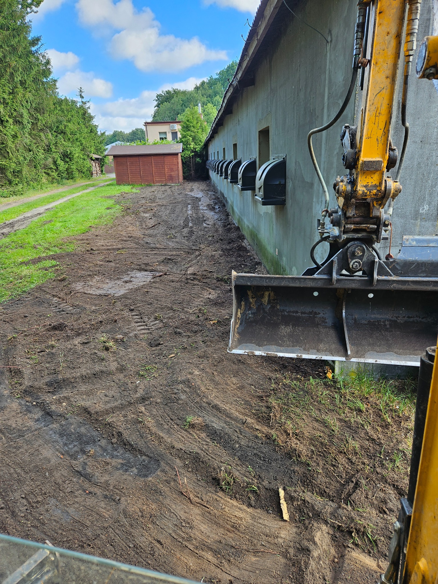 Żółta koparka z łyżką przy budynku z szarym tynkiem i rzędem czarnych okien, przygotowująca teren podłoża z widocznymi śladami opon i błotem.