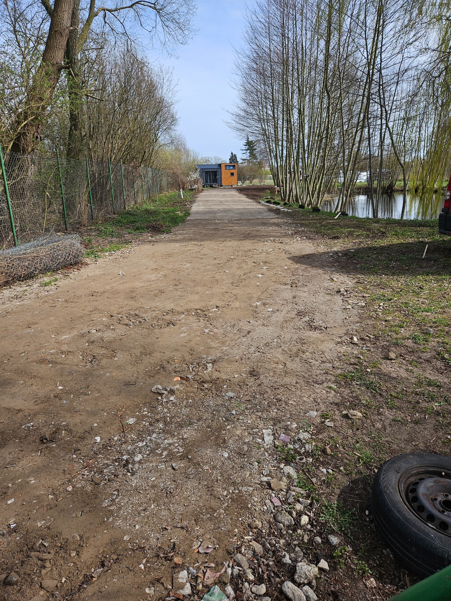 Długa, gruntowa droga w perspektywie, prowadząca do małego, nowoczesnego domu. Po lewej stronie siatka ogrodzeniowa i zwoje drutu, po prawej rzadki szpaler drzew i staw. Widoczne nierówności terenu...