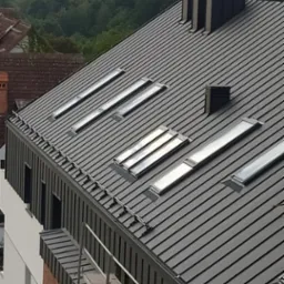 Nowoczesny, grafitowy dach z blachy na budynku w trakcie wykończenia, widoczne okna dachowe, komin i rusztowanie, w tle panorama miasta z wieżą kościelną.