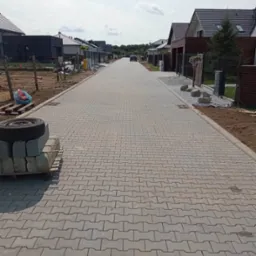 Nowa, równa droga z szarej kostki brukowej w kształcie litery H, otoczona nowo wybudowanymi domami jednorodzinnymi, z paletą kostki i oponą w lewym dolnym rogu, oraz samochodem w oddali.