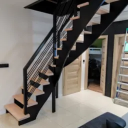 Nowoczesne schody z czarną metalową konstrukcją i jasnymi drewnianymi stopniami w jasnym wnętrzu domu.