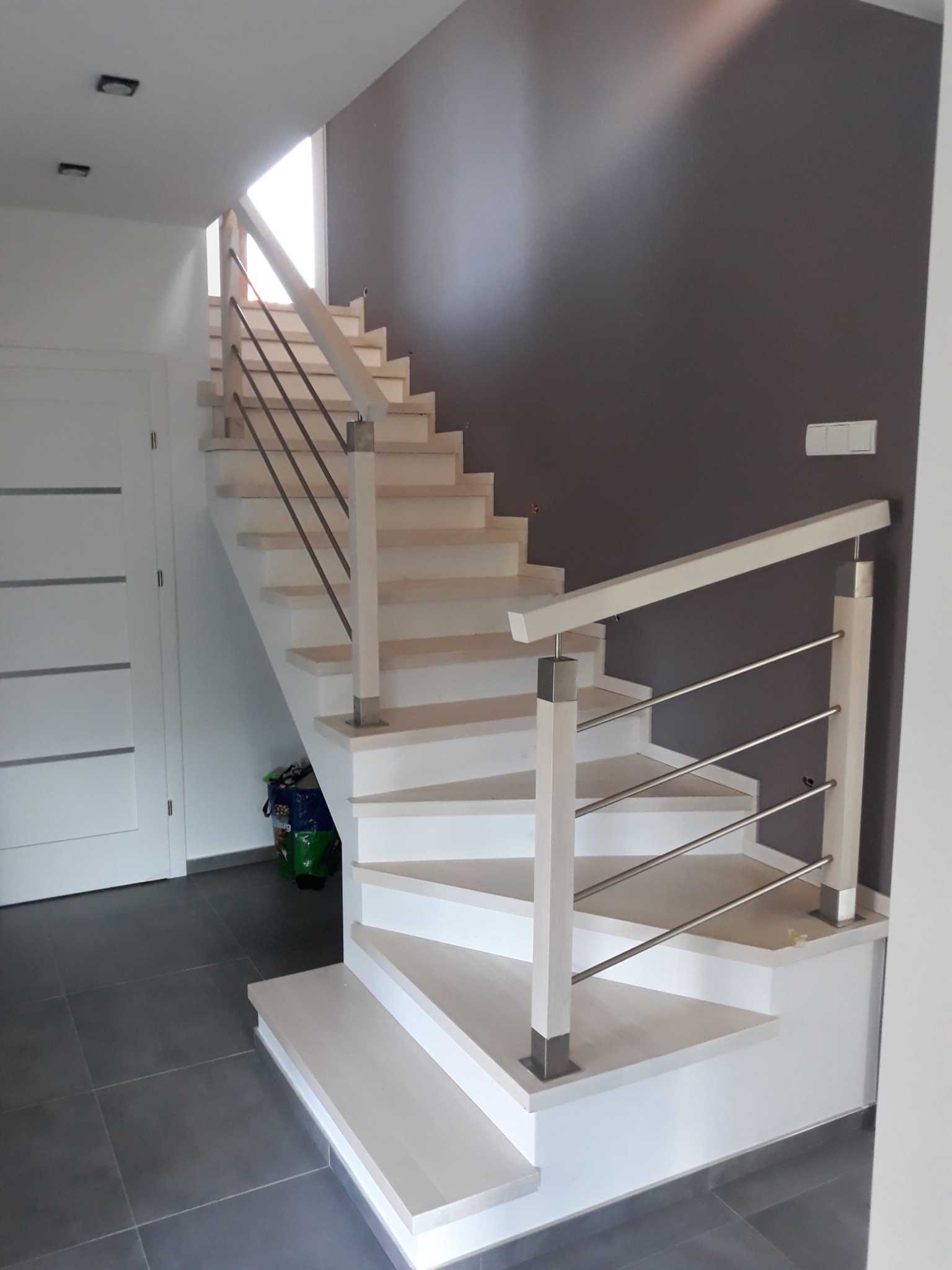 Jasne drewniane schody z metalowymi balustradami w nowoczesnym wnętrzu z szarą ścianą akcentującą.