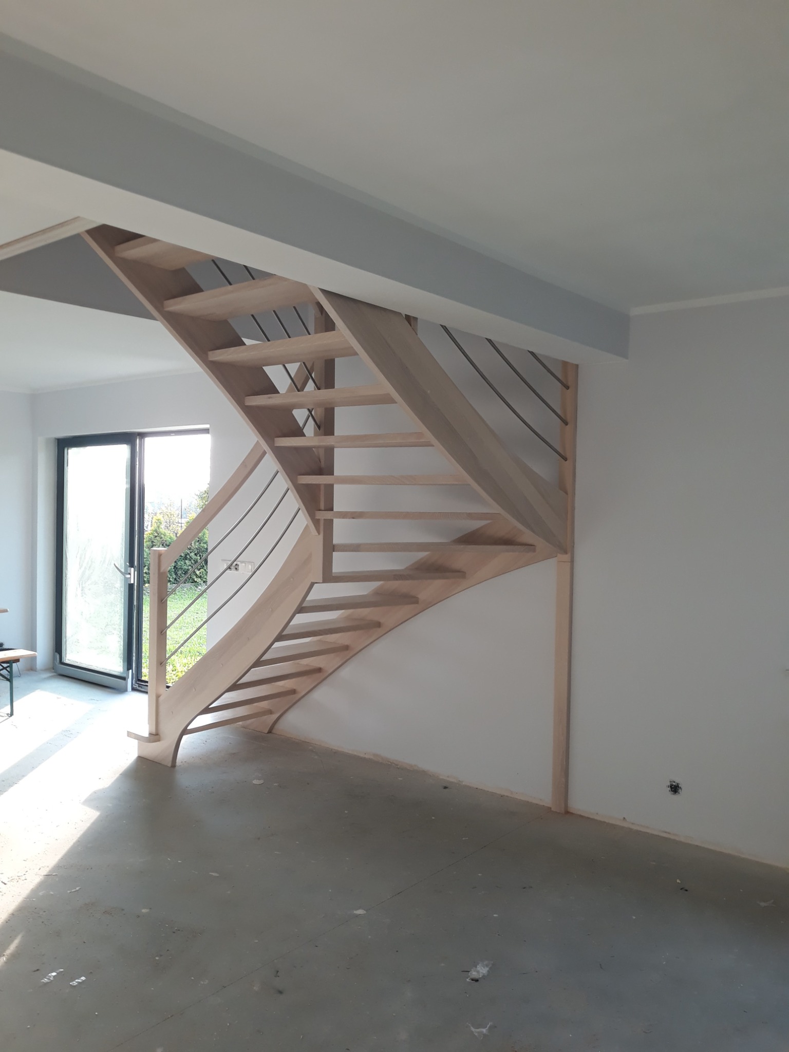 Jasne, drewniane schody z metalowymi balustradami w surowym wnętrzu, widoczne niedokończone ściany i podłoga, światło słoneczne wpada przez okno.