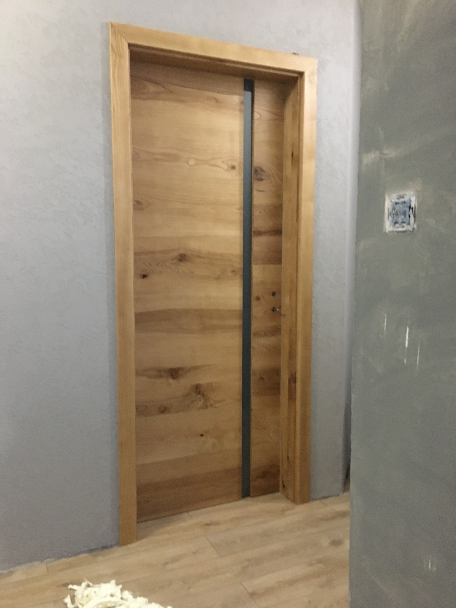 Nowoczesne drzwi z jasnego drewna z pionowym, szarym akcentem, osadzone w surowym, szarym wnętrzu z widocznymi śladami prac wykończeniowych.